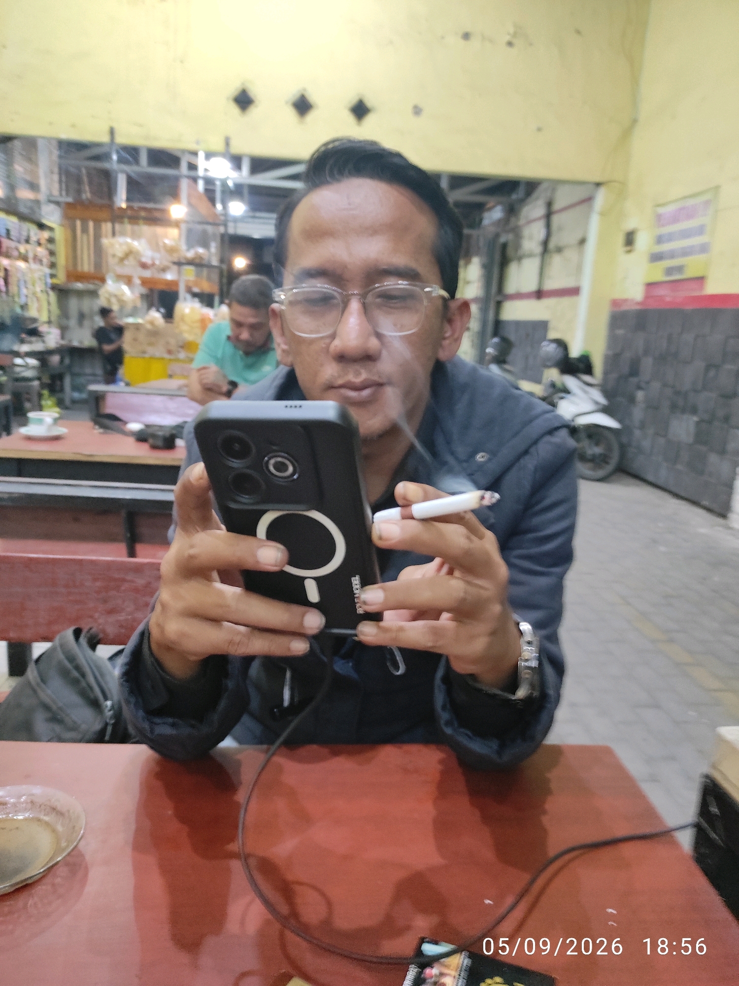 The image captures a medium shot of a man, likely in his late 20s or early 30s, sitting at a red table in what appears to be an outdoor food stall or warung in Surabaya, Indonesia. He is holding a black smartphone in his left hand and a lit cigarette in his right hand, from which smoke is emanating. He is wearing glasses and a dark-colored jacket. In the background, blurred figures and merchandise are visible, suggesting a busy commercial area. The timestamp at the bottom right corner indicates the date and time as "05/09/2026 18:56".