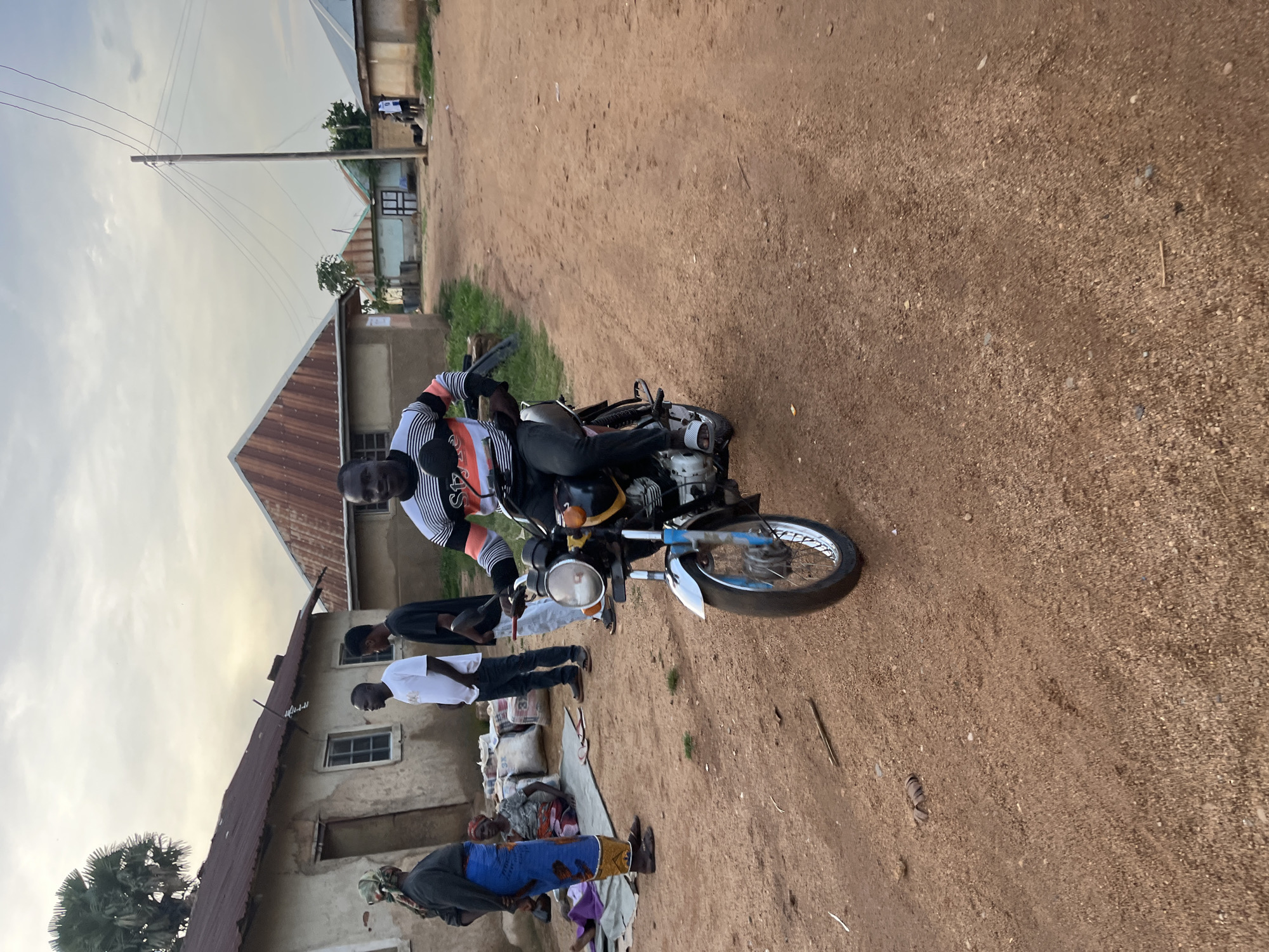 This image, oriented vertically, depicts a lively street scene in what appears to be a residential or semi-urban area, consistent with the given location context of Jos, Nigeria.
In the foreground, a man sits astride a silver and black motorcycle, prominently featuring a round headlight and blue accents on its front fender. He wears a distinctive long-sleeved shirt with black, white, and orange stripes, and dark trousers. His gaze is directed towards the viewer. The ground beneath the motorcycle and extending throughout the scene is an unpaved, reddish-brown dirt road, dotted with small stones.
Along the left side of the image, simple single-story buildings with corrugated metal roofs line the roadside. These structures have worn, light-colored walls and small windows. Several other individuals are present: two men stand casually behind the motorcycle, one in a white t-shirt and dark pants, the other in a black shirt. Further down the road, closer to the buildings, a woman is seated on the ground next to white sacks, suggesting a market stall or goods for sale. Another woman, wearing a blue patterned wrapper and a dark top, stands nearby. In the background, a tall wooden utility pole with multiple wires extends into the sky, and distant figures are faintly visible. A palm tree is also visible near the buildings.
The sky, visible on the right side of the vertically oriented image, is bright but soft, indicating either late afternoon or early evening light, with some scattered clouds. The scene conveys a sense of everyday activity in a local community. No visible text is discernible on any objects or clothing.