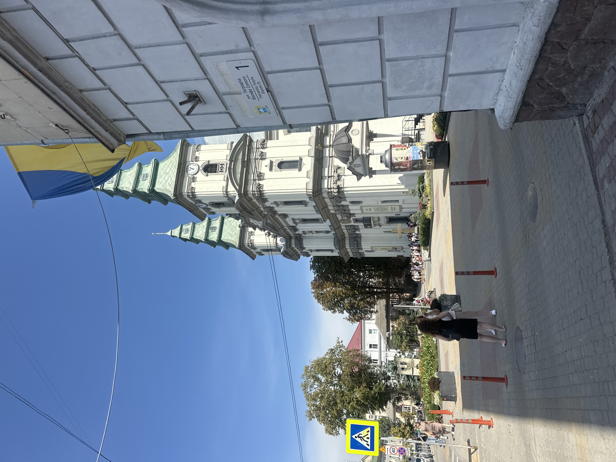 The image shows a street scene in Ternopil, Ukraine. A prominent church with two green spires and a clock tower is visible on the right side of the frame. A Ukrainian flag is hanging from a pole in front of the church. In the foreground, a white wall with brick detailing is on the right, and a street with cobblestone paving is on the left. A pedestrian crossing sign and other traffic signs are present on the left side of the street. Several people are walking on the sidewalk and across the street. The weather is sunny with a clear blue sky.