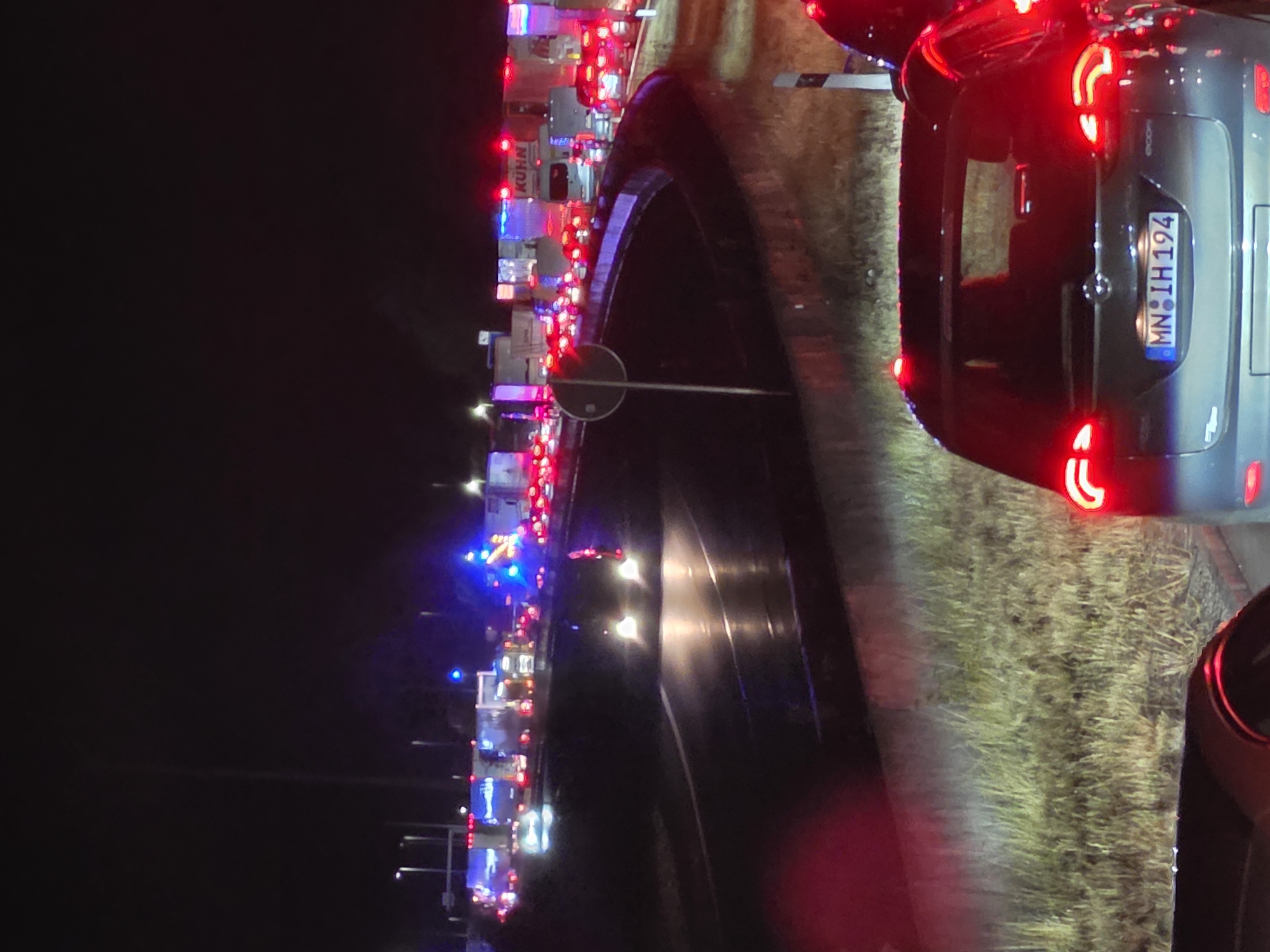 The image depicts a nighttime scene of a traffic jam on a highway, likely in or near Uettingen, Germany, evidenced by the German license plate "MN IH 194" on the car in the foreground. Several lanes of vehicles, including trucks and cars, are stationary. Emergency lights, flashing blue and red, are visible on some vehicles, suggesting a potential accident or road closure ahead. The road surface appears wet, reflecting the vehicle lights. The surrounding environment includes grassy verges and roadside vegetation. A circular sign with a white symbol on a dark background is present on a pole in the mid-ground. The overall atmosphere is dark, illuminated primarily by the artificial lights of the vehicles and emergency services.
