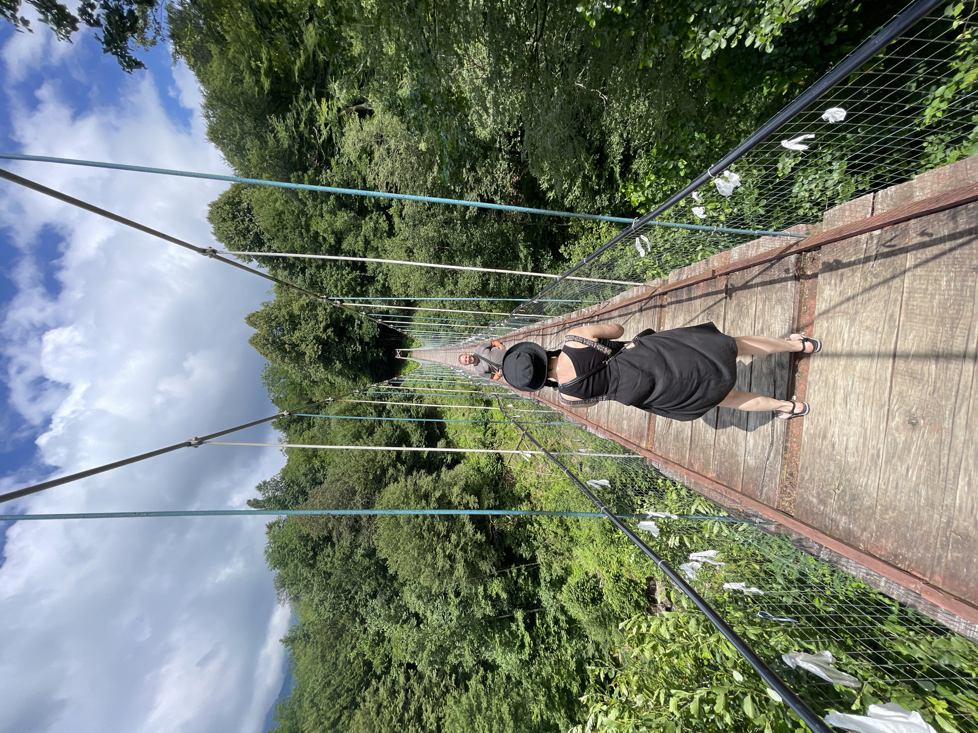 In Tqwarchal, Abkhazia, two individuals are captured from behind walking across a suspension bridge. The bridge, constructed with wooden planks and metal supports, is surrounded by lush green forestation. The sky above is partly cloudy, with patches of blue visible. The woman in the foreground wears a black hat and a black dress, while the man further down the bridge wears a grey t-shirt. The bridge has a protective metal railing on either side, adorned with white fabric scraps. The perspective is from behind the woman, looking towards the man and the continuation of the bridge into the dense forest.
