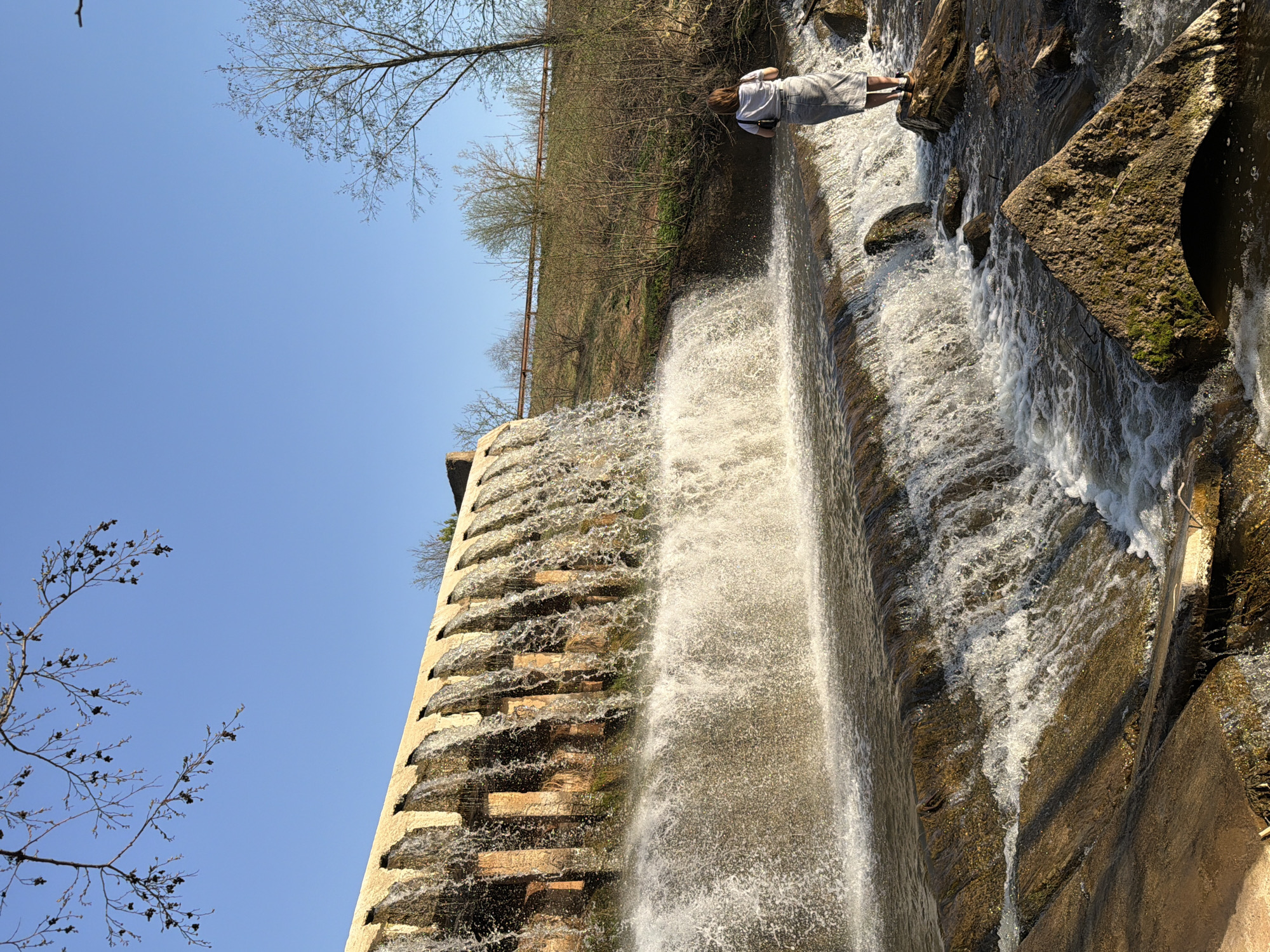 The image shows a stepped dam or waterfall, presumably in or near the city of Kamennoye, Russia. The sky is a clear blue with bare tree branches visible in the upper left corner and along the top edge of the image. A person is standing at the top of the dam, near the edge where the water begins to flow. The dam structure appears to be made of stone or concrete with a stepped design, allowing the water to cascade down. The water is turbulent and frothy as it flows over the steps. The surrounding environment consists of some sparse vegetation on the embankment.