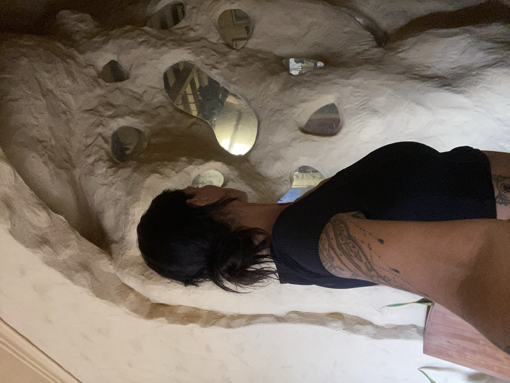 The image captures a woman standing with her back to the camera, facing a textured, beige-colored wall with irregular holes that reflect light. She has dark hair pulled back into a bun and is wearing a black, short-sleeved top. A tattoo is visible on her left arm. To the left of the woman, a wooden object and some greenery can be seen. The scene appears to be indoors, possibly within a unique architectural structure. The presence of reflections within the holes suggests they might be openings or embedded mirrors. The overall impression is an artistic or unconventional interior space. The location is not explicitly identifiable from the visual information provided.