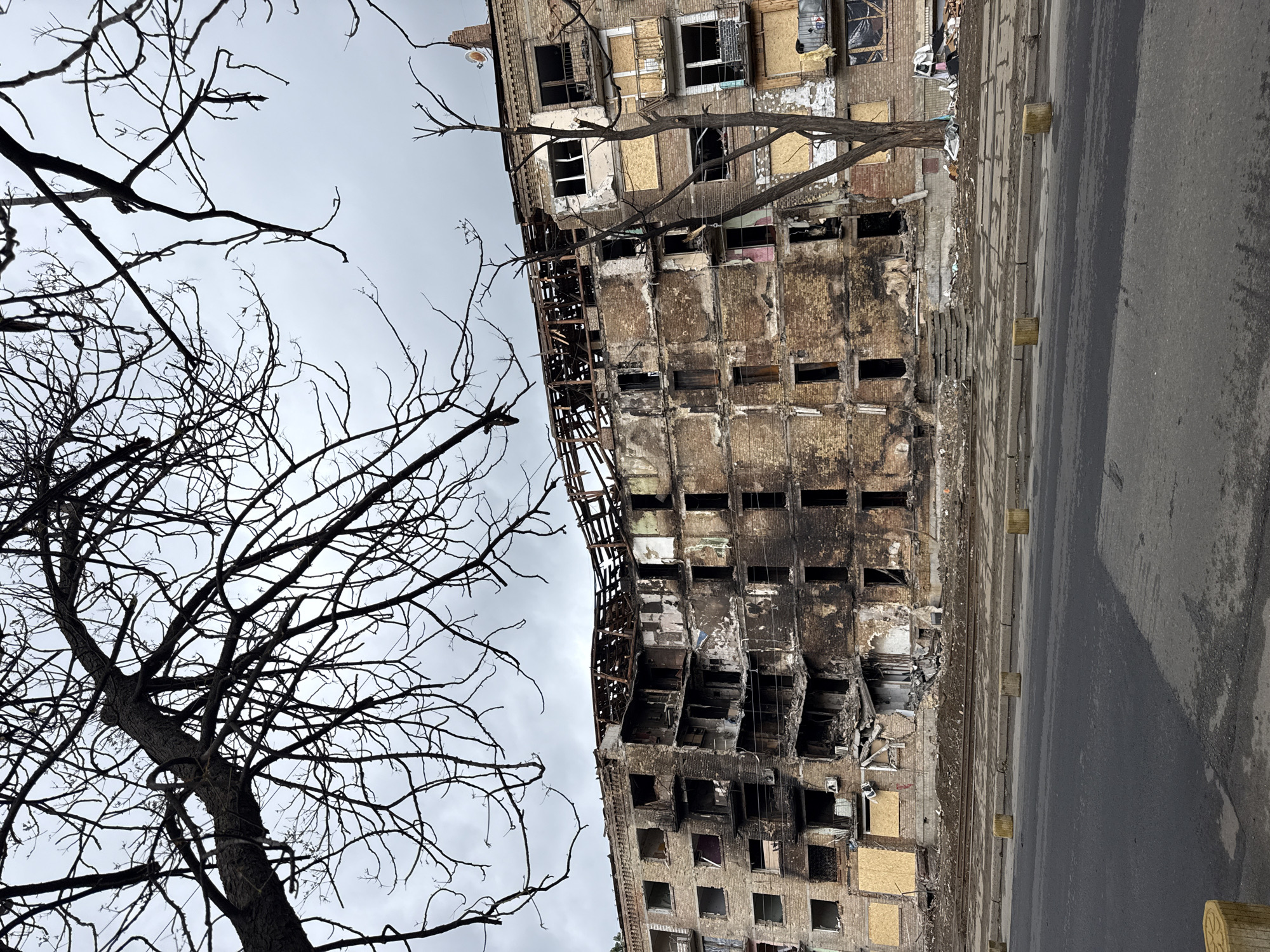 The image shows a heavily damaged building in Kyiv, Ukraine. The building appears to be a multi-story residential structure, with significant fire and structural damage. Many windows are boarded up or broken. The facade is charred and crumbling. A leafless tree stands in the foreground, adding to the starkness of the scene. A road runs along the right side of the building. The sky is overcast. There are no people visible in the image.