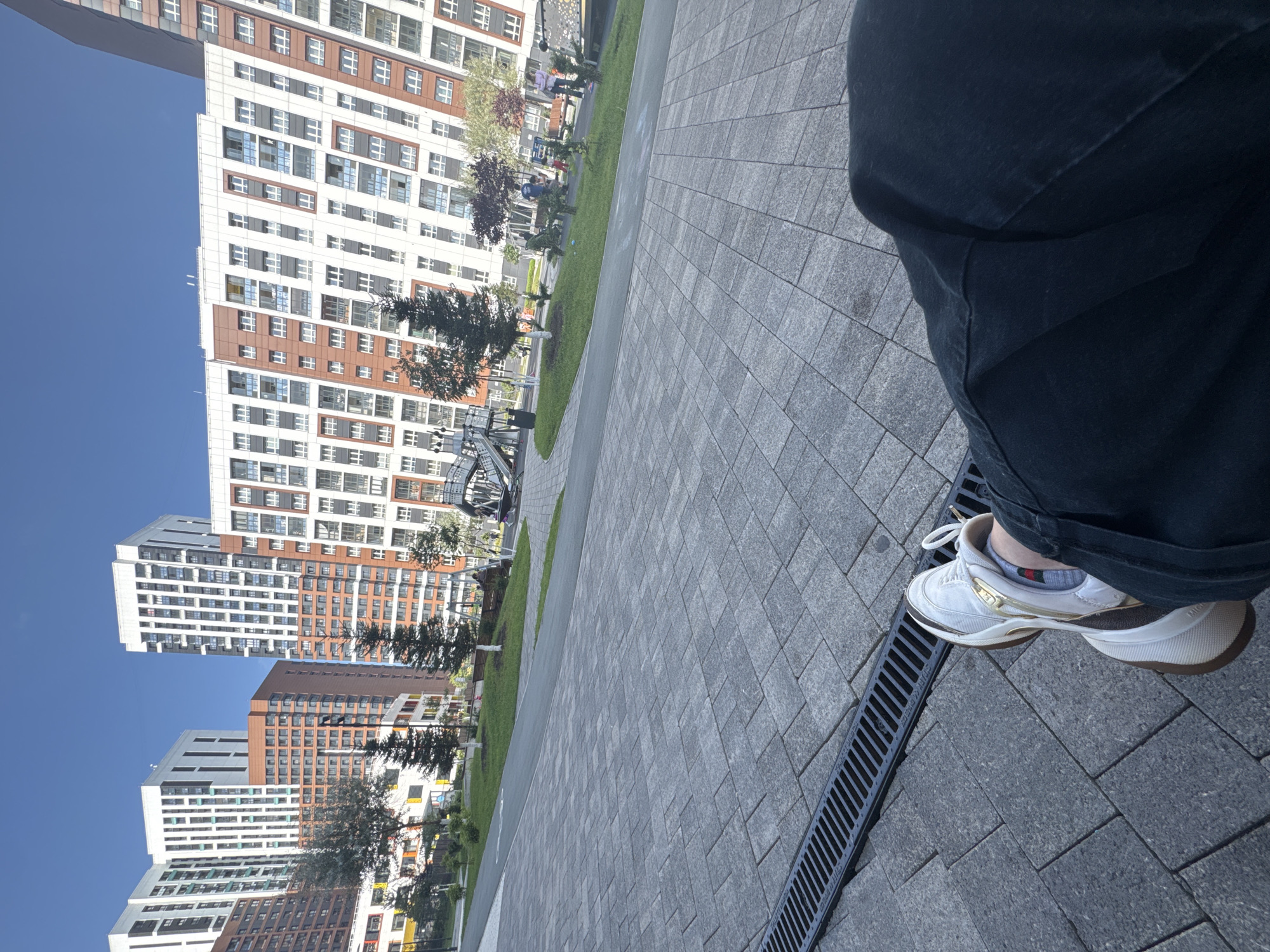 The image shows a person's legs and feet, wearing dark pants and white sneakers, sitting on a paved area in a courtyard with a drainage grate in the foreground. In the background, there are several modern, multi-story apartment buildings with white and orange facades and numerous windows. Trees and a playground are visible in front of the buildings. The sky is clear and blue. The scene appears to be a residential area in the city of Novosibirsk, Russia, likely in a newly developed neighborhood.