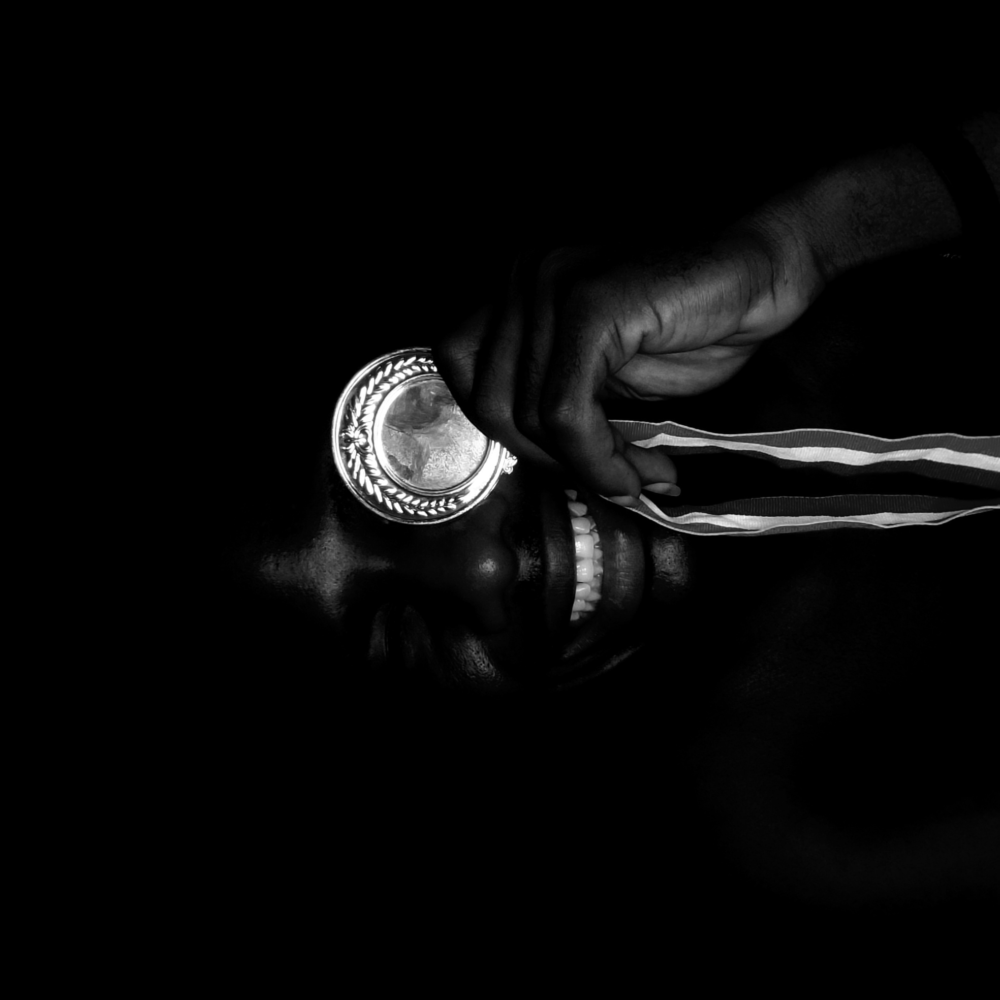 A black and white, close-up, angled, high-contrast image shows a person smiling while holding a medal near their eye. The person has dark skin and visible white teeth. A hand holds a silver medal with a decorative edge. The medal's ribbon is partially visible, displaying a striped pattern. The background is dark, obscuring details of the environment. It is not possible to confirm the location.