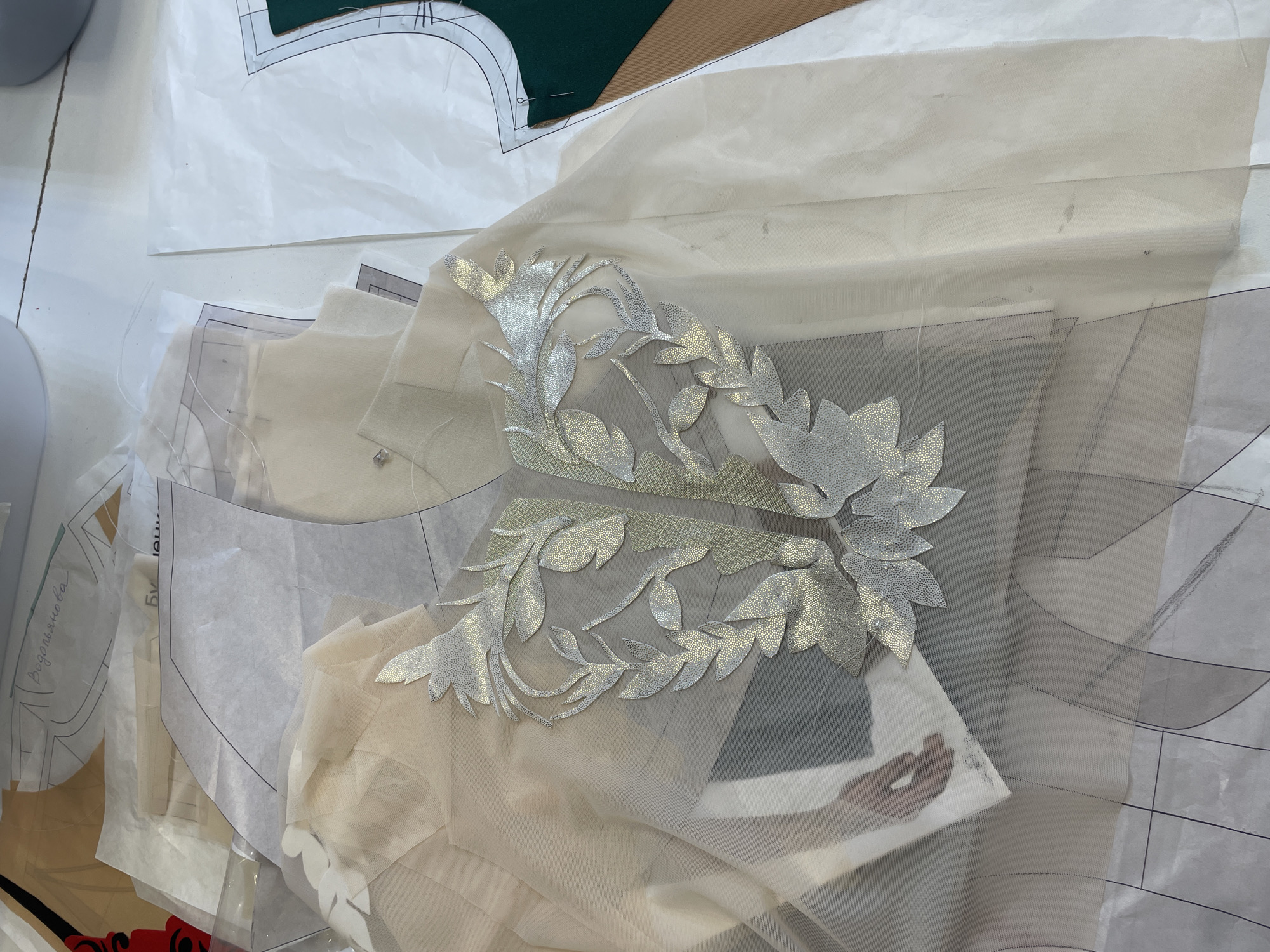 In an overhead shot of a cluttered workbench, a sheer, cream-colored fabric lies spread out, adorned with a silver, leaf-like appliqué design. Beneath the fabric, several paper sewing patterns are visible, some with handwritten notes in Russian, including the name "Водопьянова" (Vodopyanova). To the right, a green fabric piece is pinned to a pattern. The overall scene suggests a sewing or design studio, possibly in the city of Unknown, Unknown, where garments are in the process of being created.