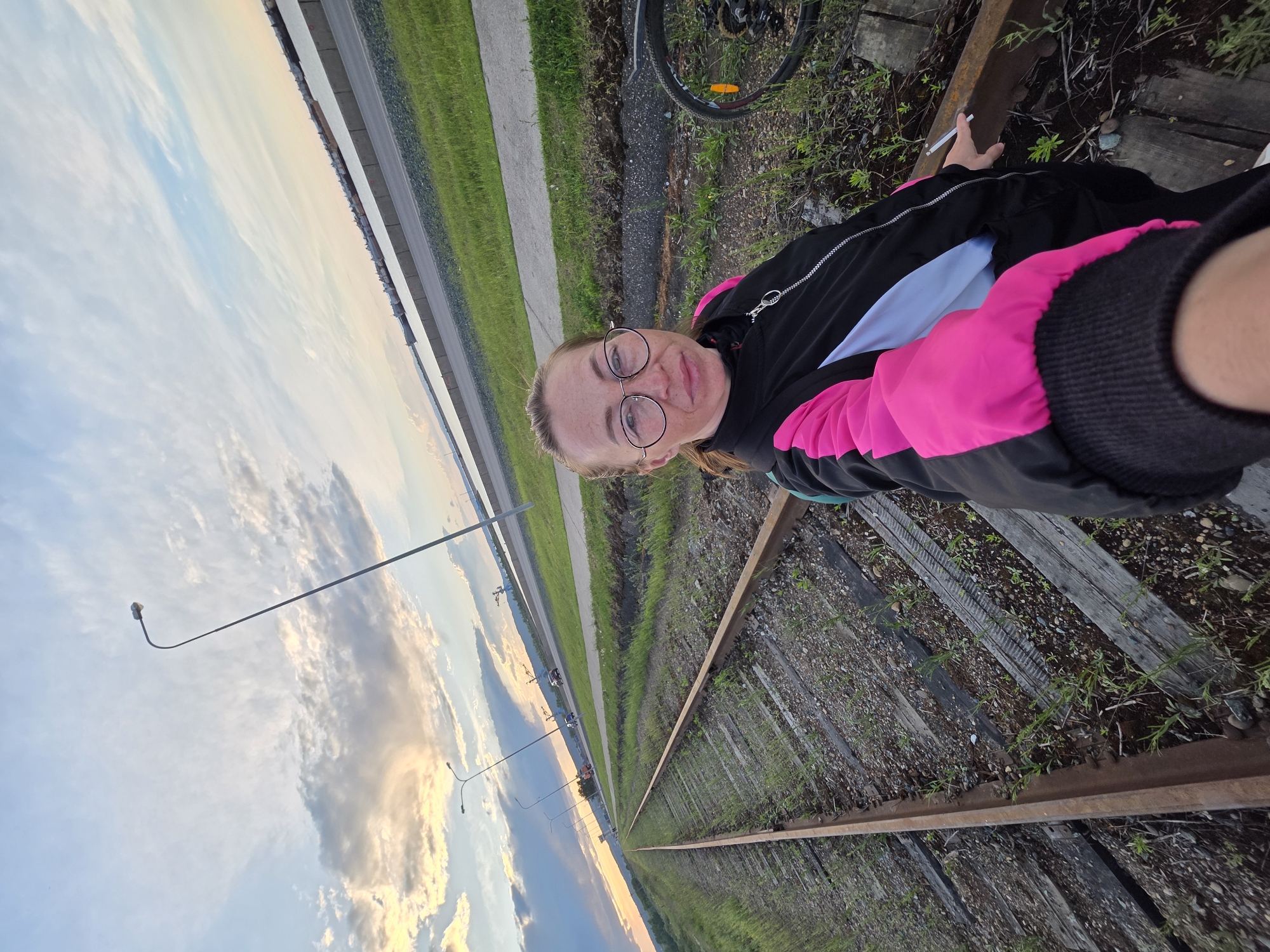 A woman with round glasses poses for a selfie on railroad tracks. She wears a black jacket with a pink and white design. A bicycle wheel is visible in the upper right corner. In the background are streetlights, a road with cars, a grass-covered area, and a cloudy sky. The scene appears to be taken in the city of Chaikovsky, Russia.