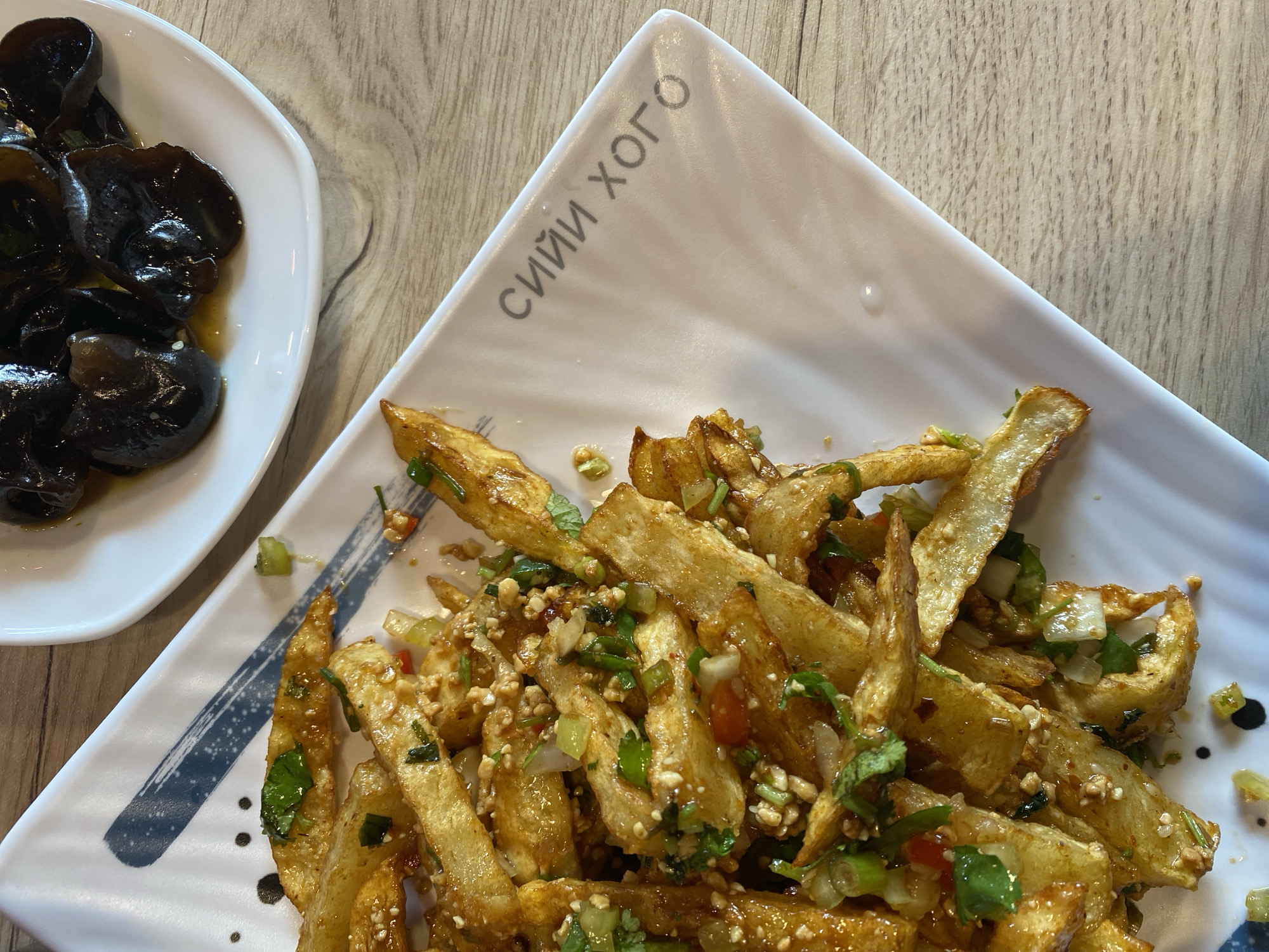This image shows two white ceramic plates on a light-colored wooden table. The plate in the foreground is rectangular with a patterned rim and contains a pile of golden-brown french fries coated in a glistening sauce and garnished with chopped green herbs, red chili flakes, and minced peanuts. The plate in the background is oval and holds a serving of dark, glossy ingredients, possibly wood ear mushrooms in a sauce. The overall impression is of a restaurant setting, likely in Saint Petersburg, Russia, given the visible text on the foreground plate, which appears to be in Cyrillic script and reads "СИЙИ ХОГО." The lighting suggests an indoor environment, possibly a restaurant with natural or ambient light.