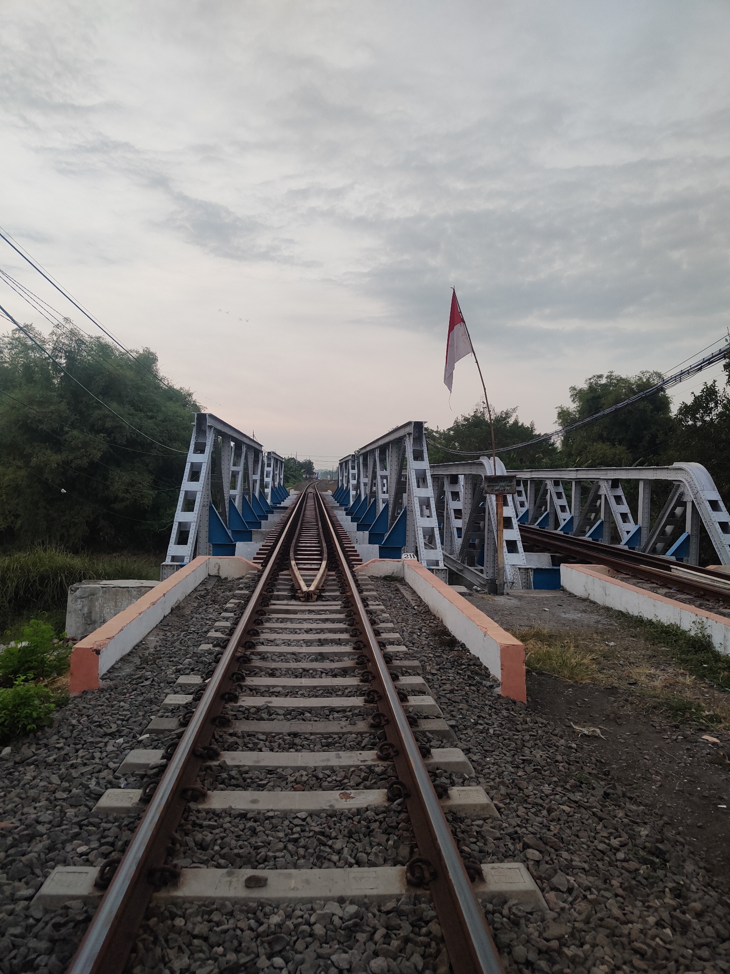 An outdoor, low-angle, medium shot shows a pair of railroad tracks running straight ahead towards a steel truss bridge. The bridge has blue accents and a red and white flag is waving from a pole on the right side of the bridge. Lush green trees and foliage line the left side of the tracks, while thinner trees and vegetation are visible on the right. The sky is overcast with soft clouds. The railroad tracks are made of steel rails laid on concrete sleepers and ballast. There are no people or vehicles visible in the image. The scene appears to be in Kauman, Indonesia, as indicated by the Indonesian flag.