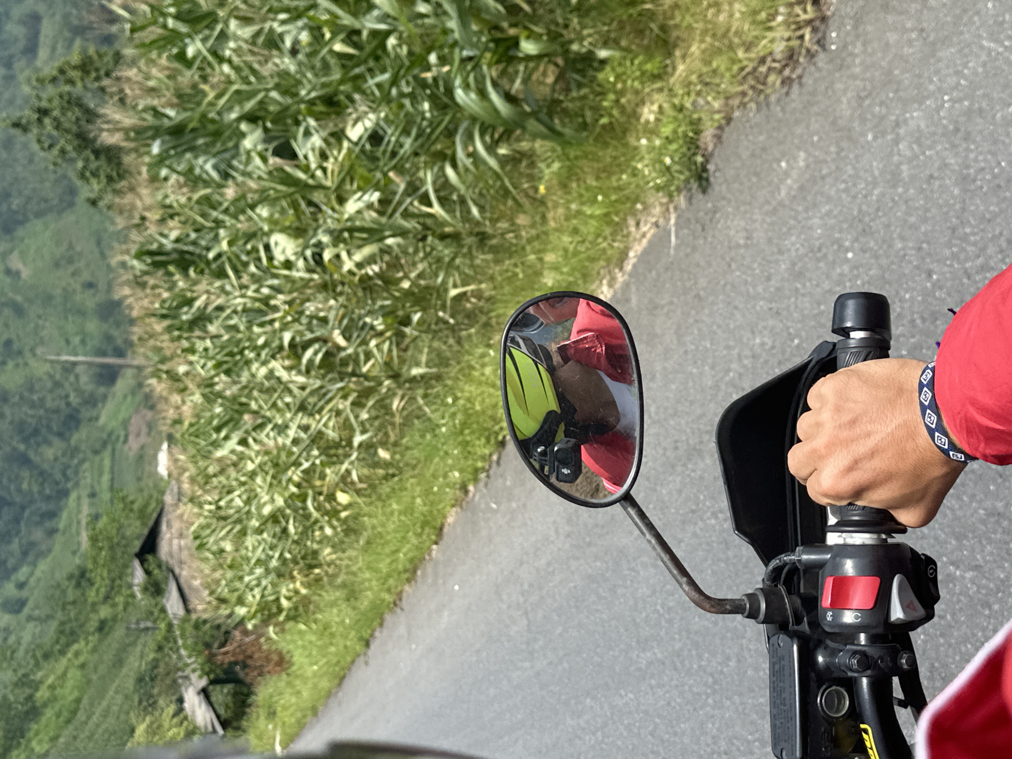 This first-person perspective image captures a scene from a motorcycle journey through a verdant, rural landscape, consistent with Minh Sơn Commune, Vietnam. The view looks forward from the rider's position, showing the right handlebar grip held by a hand adorned with a dark bracelet featuring white geometric patterns. The rider wears a red long-sleeved shirt.
The paved road runs alongside a steep embankment densely covered in lush, tall green vegetation, possibly agricultural crops like corn. In the distance, rolling green hills or mountains extend under a bright daytime sky, indicating sunny weather. A small, dark structure or shack is partially visible nestled among the greenery on the left side of the road.
The motorcycle's left rearview mirror offers a reflection of a passenger or second rider, who is also wearing a red shirt and a bright yellow-green vest or backpack. A small black action camera is mounted and visible within this reflection. The handlebar controls include a prominent red button and a gray switch, with indistinct symbols (one resembling a 'C' with a partial circle, another a triangle) visible nearby. The image is oriented sideways, suggesting a dynamic capture during the ride.