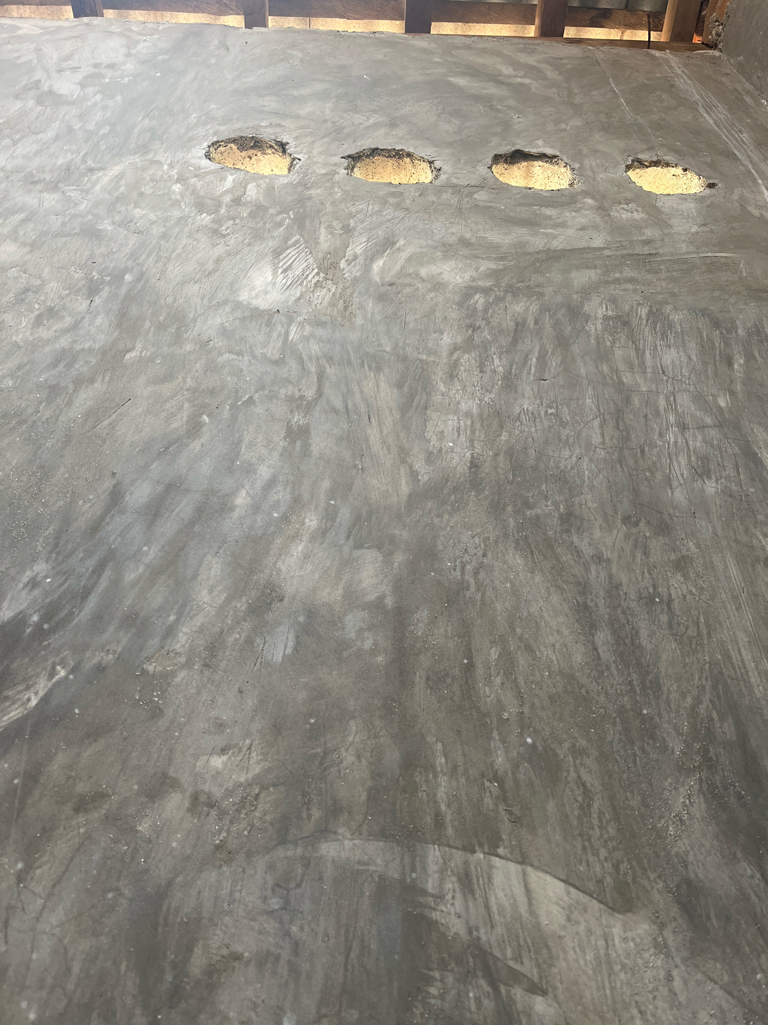 The image shows a concrete floor with four oval-shaped openings, each appearing to be filled with a yellowish, textured material, possibly straw or hay. The concrete surface has a rough, unfinished texture with visible brush strokes and some lighter-colored splotches. In the background, the top edge of the image shows wooden beams, suggesting an unfinished structure or ceiling. The scene appears to be outdoors or in an open-air construction site. There are no people or animals visible in the image. The lighting suggests it is daytime, with the light source coming from above and slightly to the left, casting faint shadows on the concrete. The location is specified as Kebumen, Indonesia.