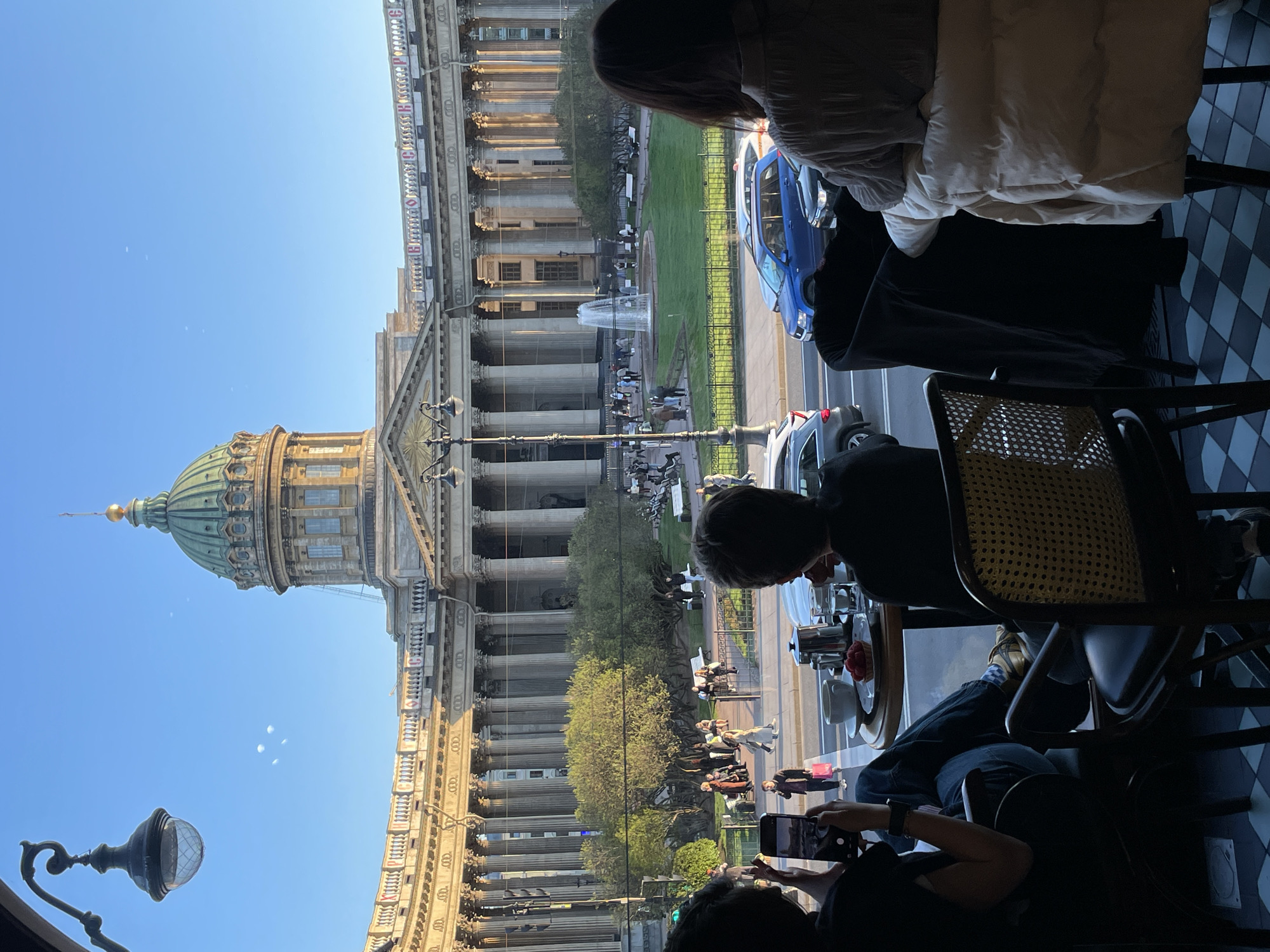 The image shows the Kazan Cathedral in Saint Petersburg, Russia, viewed from inside a cafe. The cathedral's facade, including its colonnades and dome, are visible through the window. People are visible inside the cafe, sitting at tables. One person is taking a picture of the cathedral with their phone. A blue car is parked on the street outside. A fountain is visible across from the cathedral. Other people are visible in the street and the park in front of the cathedral.