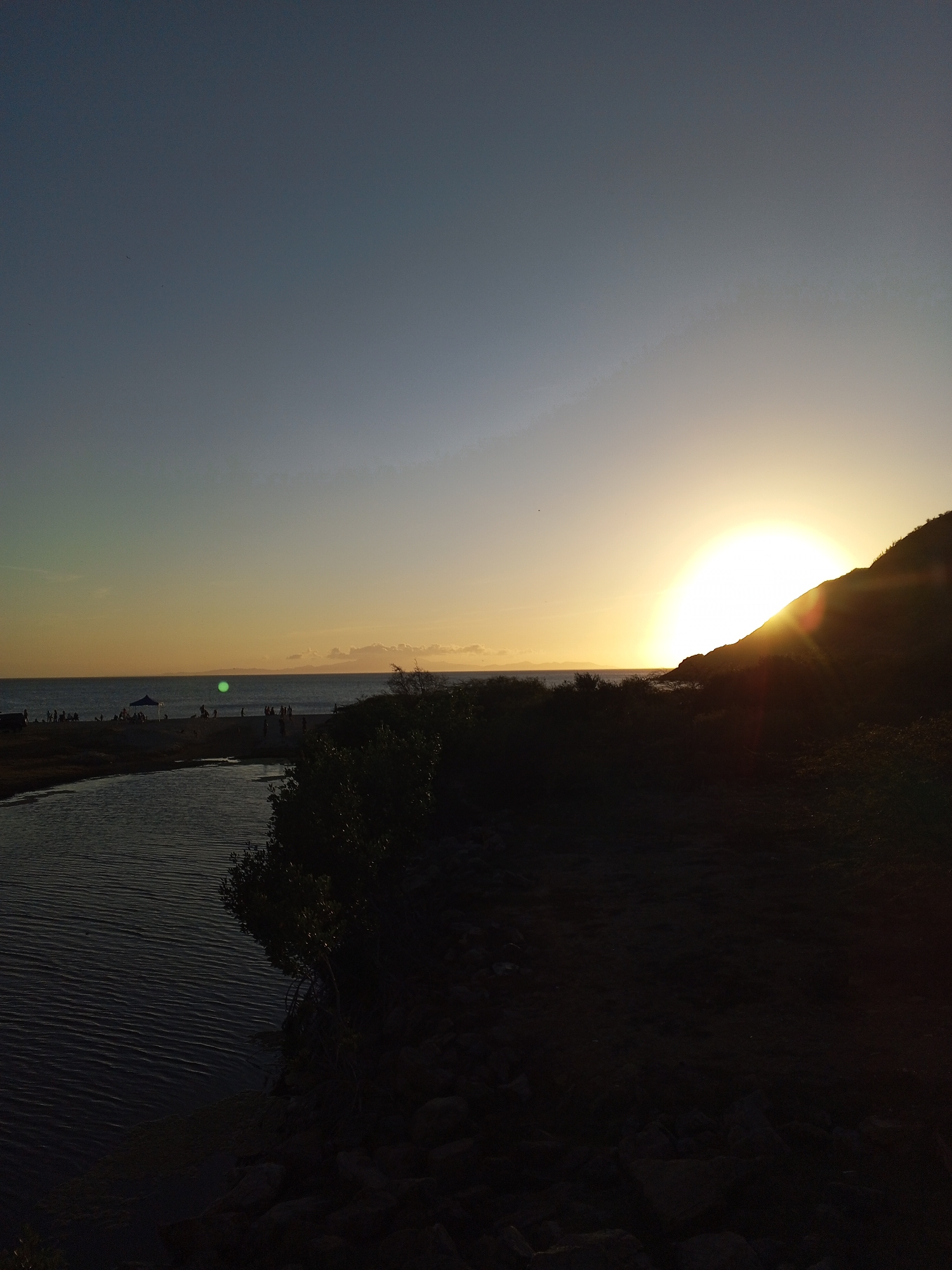 The image shows a sunset in Juan Griego, Venezuela. A bright sun is setting over a hill on the right. Below the hill, there is dark vegetation and a rocky foreground. To the left is a body of water, possibly a lagoon or river, leading out to the sea. On the beach near the sea, there are people and a blue tent visible in the distance. The sky is a gradient of blue to yellow-orange as it approaches the horizon.