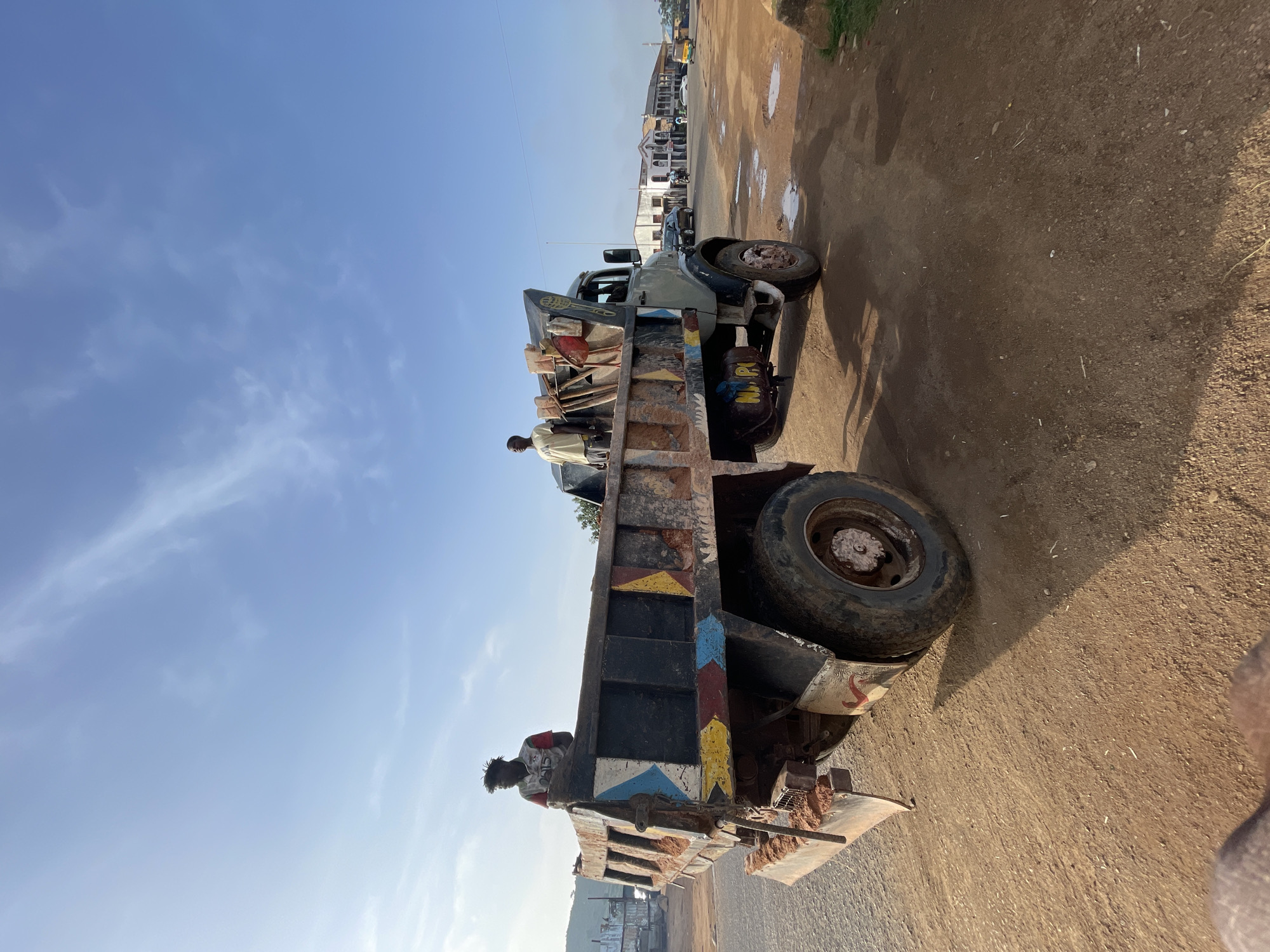 The image captures a scene on a sunny day in Jos, Nigeria, likely on a dirt road near buildings. A weathered, orange-brown dump truck, filled with soil, is the central focus. Two individuals are visible in the truck bed: one, wearing a red, white, and green shirt, stands towards the rear, while another, in a light yellow t-shirt, is positioned further forward. The truck's sides are adorned with colorful, faded geometric patterns. In the background, a street lined with buildings, some appearing commercial, stretches into the distance. Several vehicles, including a tricycle, are present on the road. The ground is predominantly dry, red earth with some scattered puddles. The sky is a clear blue with wispy white clouds.