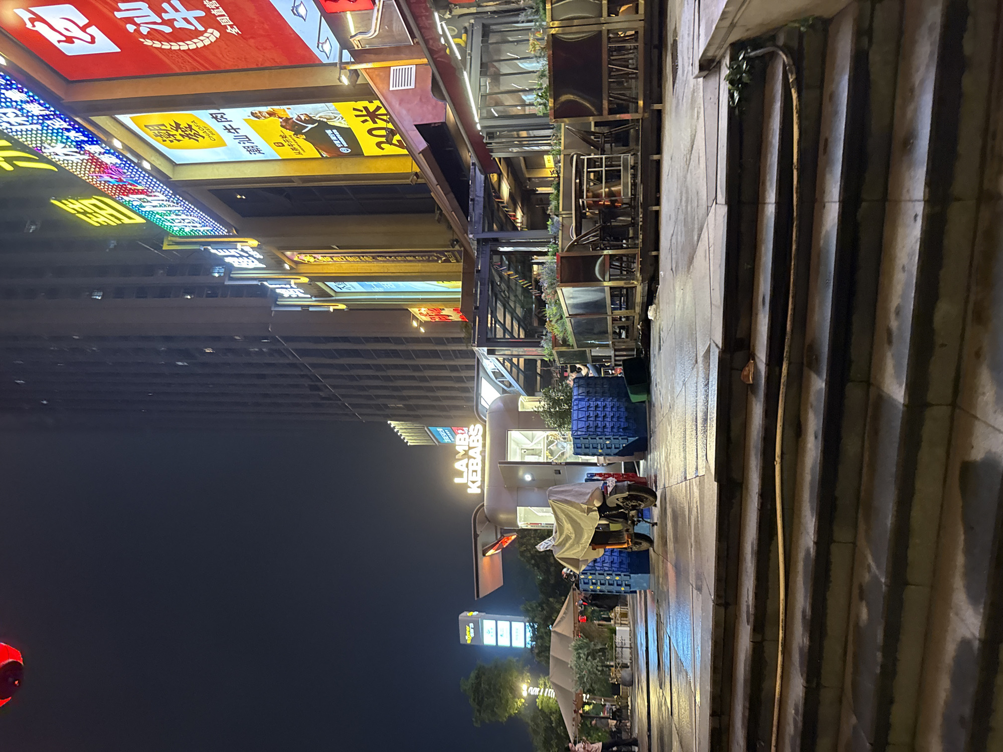 The image depicts a nighttime street scene in Haizhu District, China. The orientation of the photo is vertical, with the bottom of the image appearing on the right. The scene is illuminated by various neon signs and shop lights, suggesting a commercial area.
Prominent among the visible signs is one that reads "LAMB KEBABS" in illuminated capital letters, indicating a food establishment. Other signs display Chinese characters and colorful LED lights. In the foreground, a set of stone steps leads up from the street, and the wet pavement suggests recent rain. Several blue crates are stacked near the steps. A motorcycle covered with a tarp is parked on the sidewalk.
In the background, a tall, modern building with many windows is visible. Further down the street, more shops and businesses can be seen, also lit up with various signs. The overall atmosphere is lively, characteristic of a bustling urban environment at night. There are a few people visible, mostly blurry figures in the distance, suggesting ongoing activity, though no specific event is clearly identifiable. The time of day is clearly night, and the wet ground implies that it has rained or is still raining.