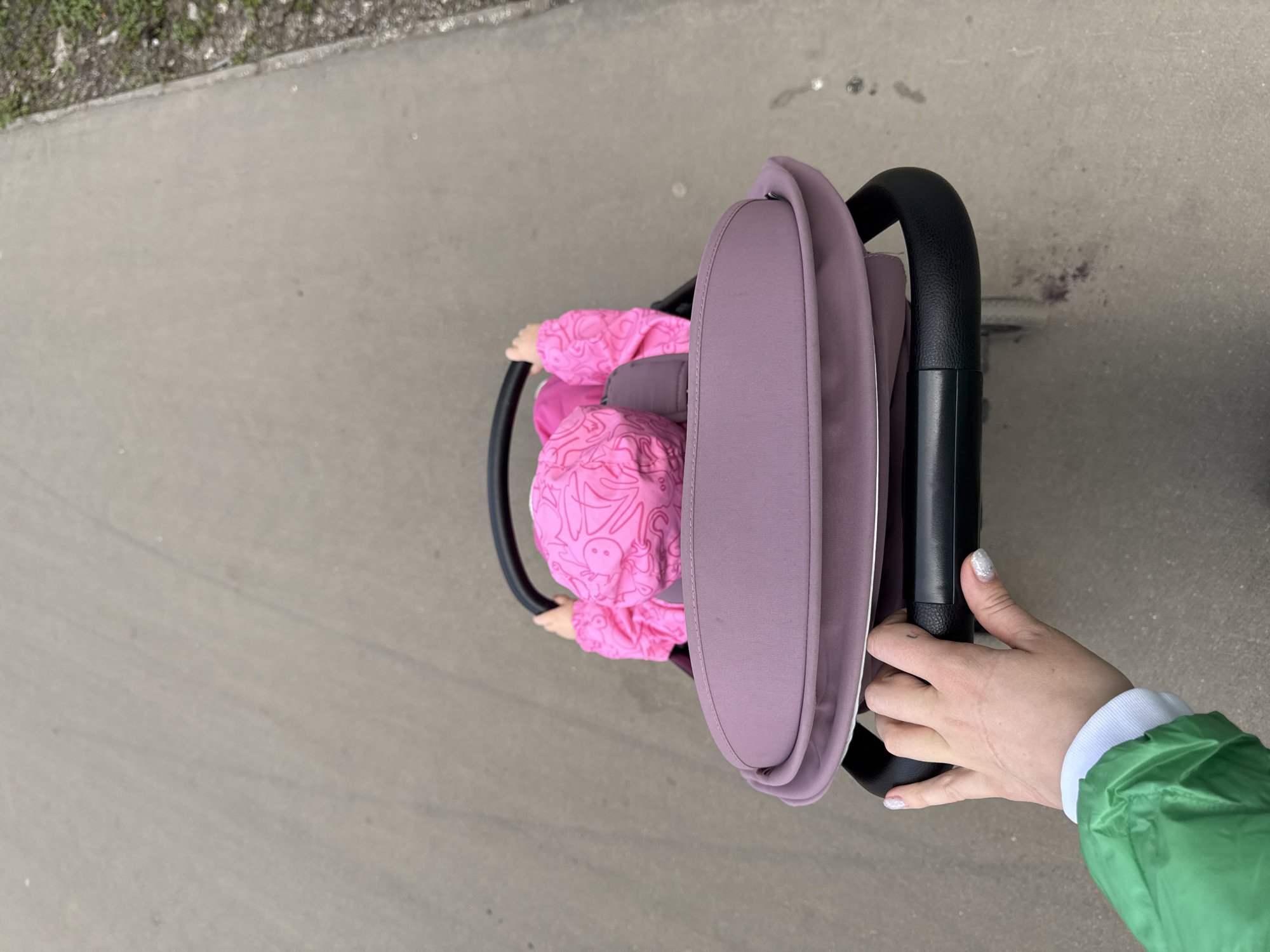 A top-down shot shows a child in a pink patterned outfit sitting in a purple stroller on a gray concrete surface, possibly a sidewalk. A person's hand wearing a green jacket is gripping the black stroller handle. The location is not identifiable as Moscow, Russia.