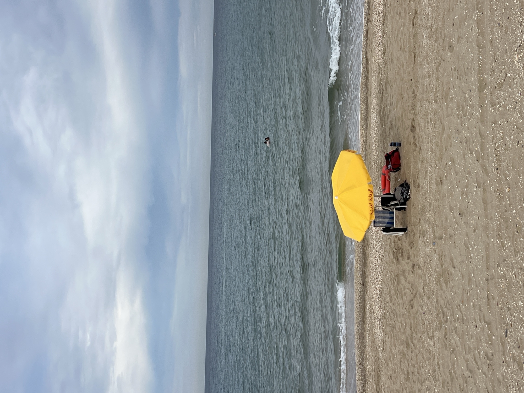 The image depicts a beach in M'Diq, Morocco. The scene is oriented with the sky and sea on the left, and the beach on the right. The sky is overcast with varying shades of gray clouds. The sea is a darker gray with visible ripples and small waves near the shoreline.
On the beach, a bright yellow beach umbrella is open, providing shade. Nearby, there is a red lifeguard buoy and a beach chair. There is also a person swimming in the ocean. The sand is light brown and composed of fine grains and shells. The scene appears calm and undisturbed.
