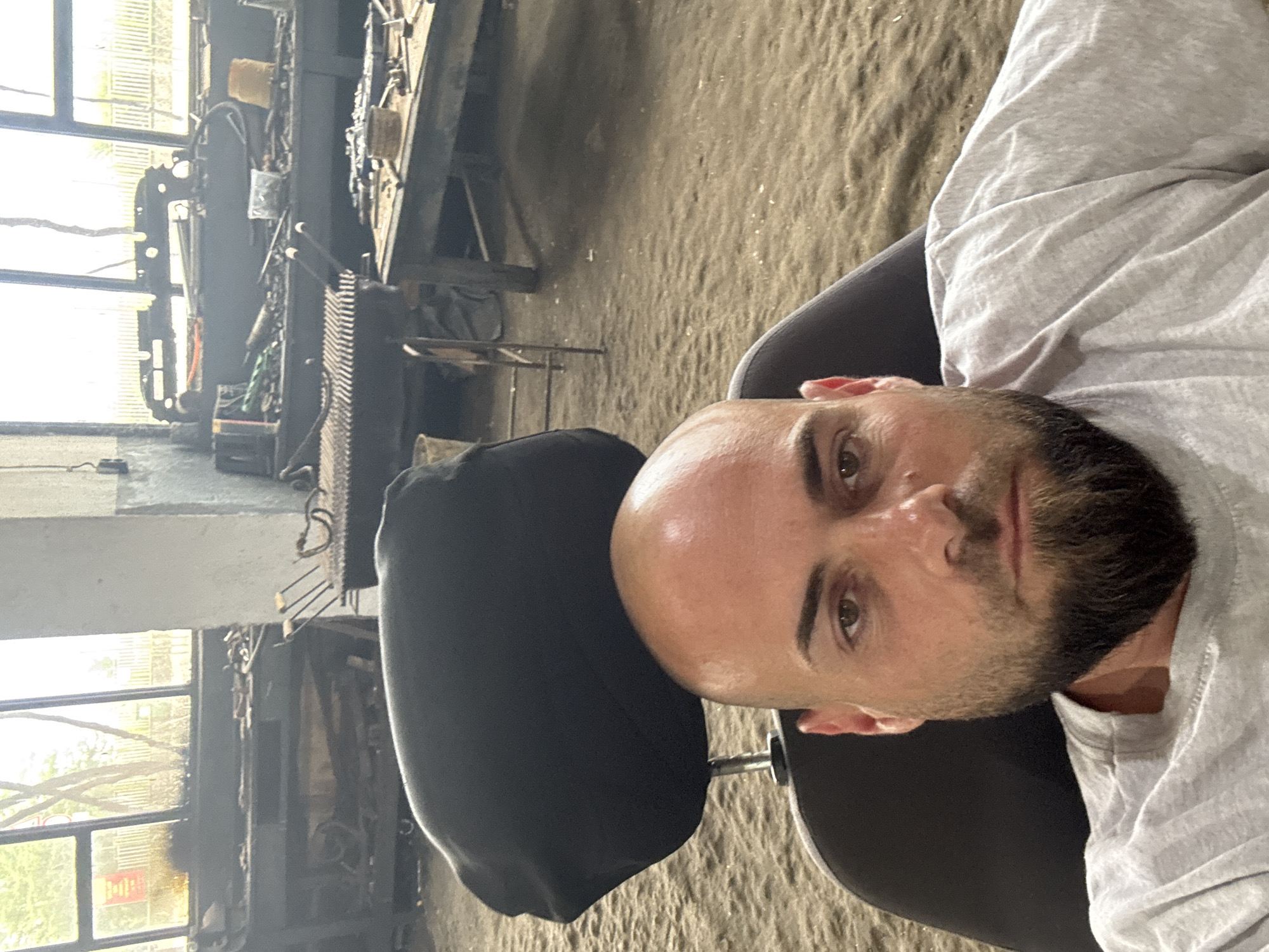 This media file features a selfie of a man taken indoors, in what appears to be an industrial workshop or garage setting in Sarnevo, Bulgaria. The original image is oriented sideways, with the man's head facing upwards and his body extending to the right.
The primary subject is a bald man with a dark beard and mustache, likely in his 30s or 40s, wearing a light grey, short-sleeved t-shirt. He has dark eyebrows and brown eyes, looking directly at the camera with a neutral expression. He appears to be seated on a black chair with a black headrest visible behind him. His forehead and scalp show some sheen, possibly from sweat or light reflection.
The background reveals a cluttered workshop environment. To the left of the man, large windows with multiple panes allow bright daylight to flood in, showing some blurred green foliage and possibly a building exterior outside. Various industrial equipment, metal structures, wires, and tools are visible throughout the background. A prominent feature behind the man's right shoulder is a ribbed, dark metal object, possibly a part of a machine or a heat exchanger. The floor appears to be an uneven, sandy-brown or light grey surface, consistent with a workshop floor.
No specific activity beyond taking a selfie is clearly depicted. The time of day is daytime, indicated by the bright light through the windows. No identifiable text is clearly legible on any objects or signs within the image, even in the cropped views. The overall atmosphere suggests a functional, possibly active, industrial space.