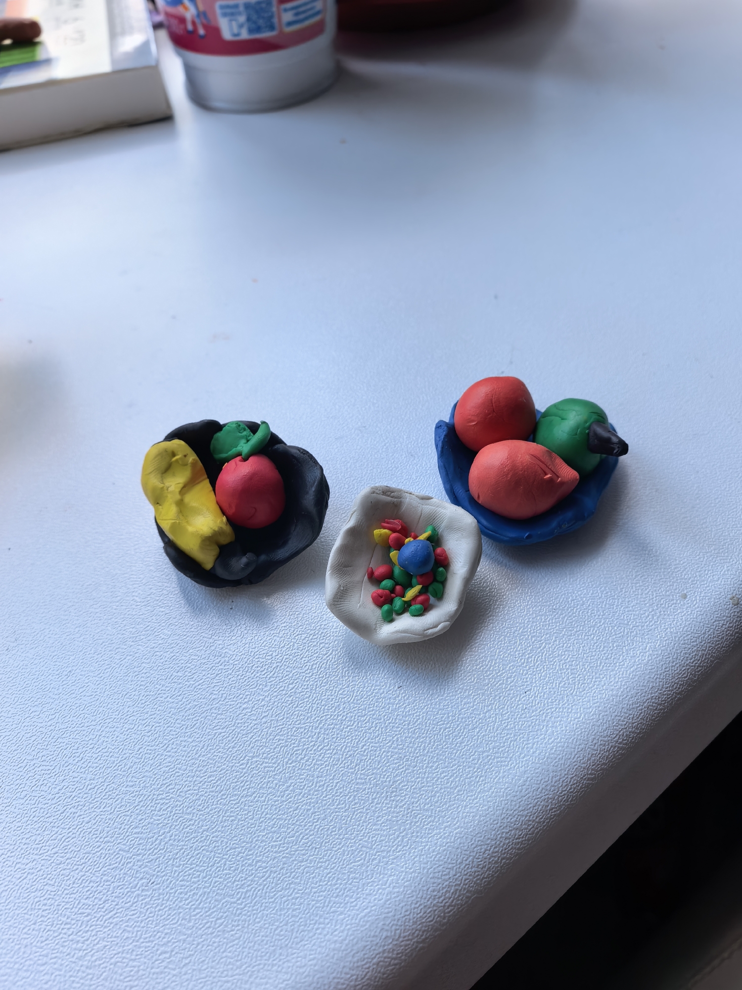 Three small clay bowls containing clay fruit sit on a light-colored, textured surface. The bowl on the left is black, containing a yellow object, a red object, and a green leaf. The bowl in the center is white and filled with small red, yellow, green, and blue clay balls. The bowl on the right is blue and contains two red objects, a green object, and a black object. The bowls are positioned diagonally across the frame. The location is unknown. There is no information to associate this scene with the city of Sterlitamak, Russia.