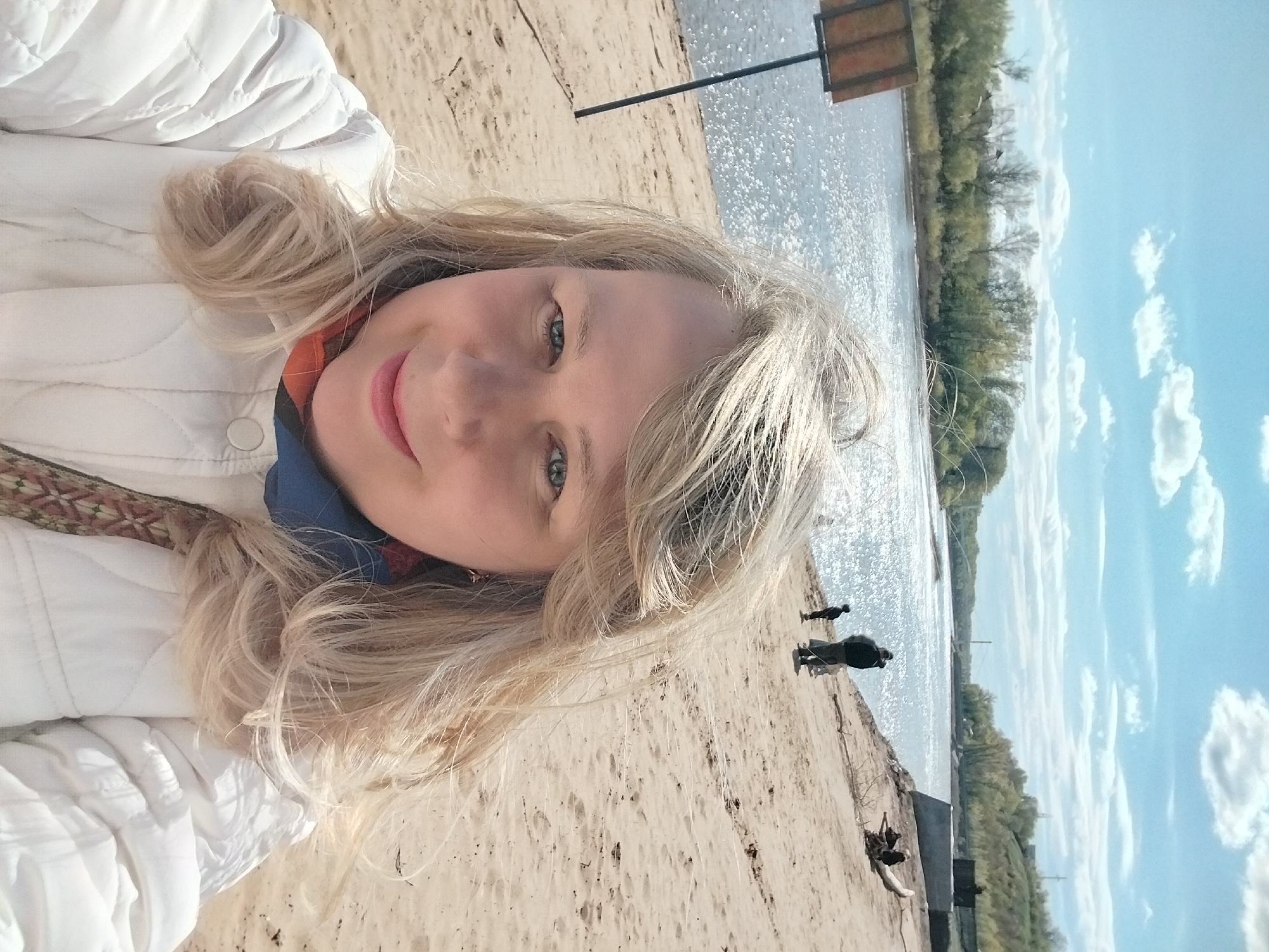 The image is a selfie taken on a sandy beach in Orenburg, Russia. The photographer, a woman with blond hair wearing a white quilted jacket, is smiling at the camera. Behind her, the beach stretches along the edge of a body of water, likely the Ural River, indicated by the glinting surface. Two people are visible further down the beach, near the water's edge. In the background, across the water, are trees and vegetation. The sky is blue with scattered white clouds.