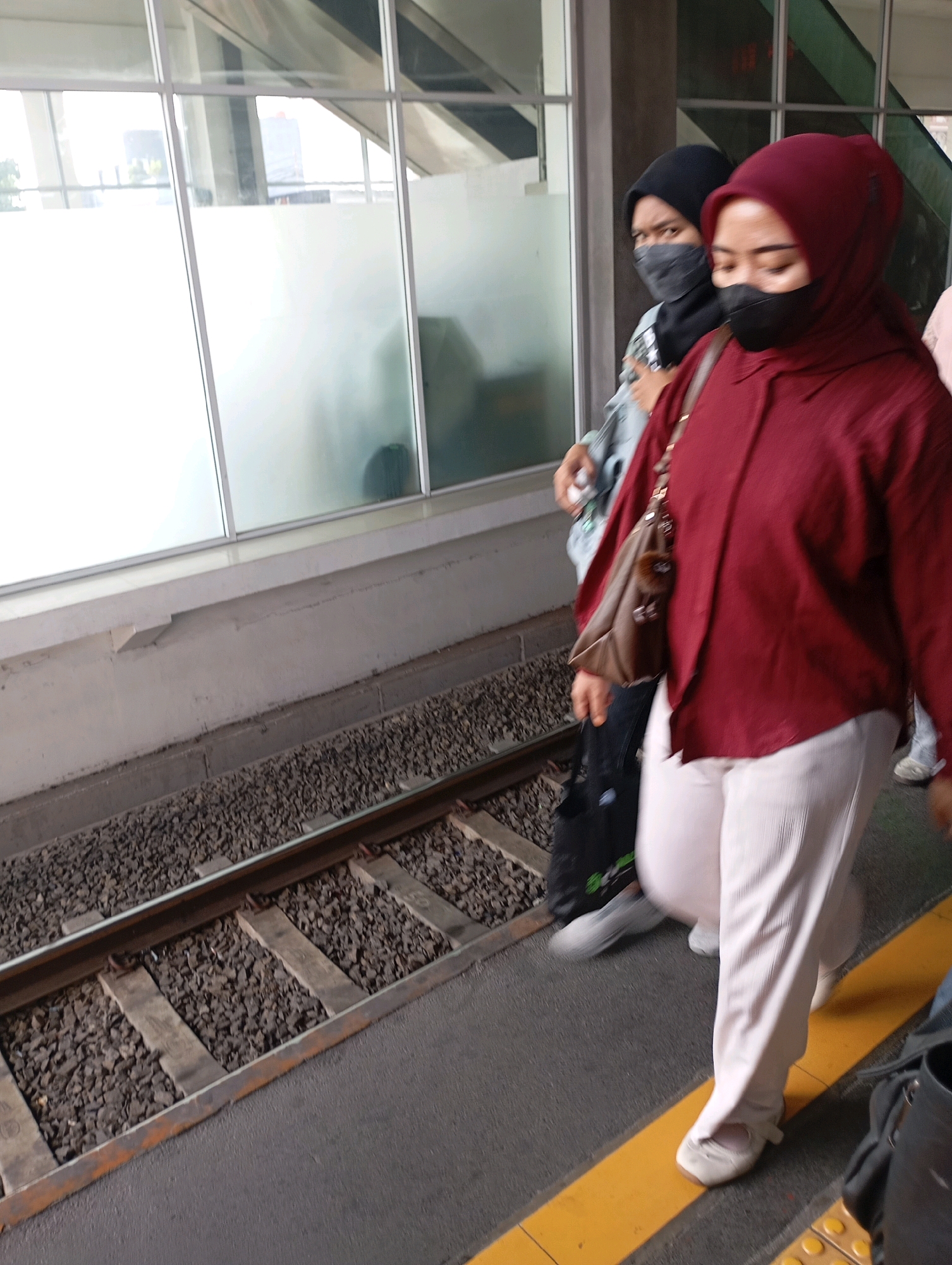 Two women wearing hijabs and masks are standing on a train platform in West Jakarta, Indonesia. The woman in the foreground is wearing a maroon top and white pants, carrying a brown shoulder bag. The woman behind her is wearing a black hijab and a grey mask, with a light blue jacket visible. They are standing near train tracks with ballast. The platform has a concrete wall and large windows with frosted and clear panels.