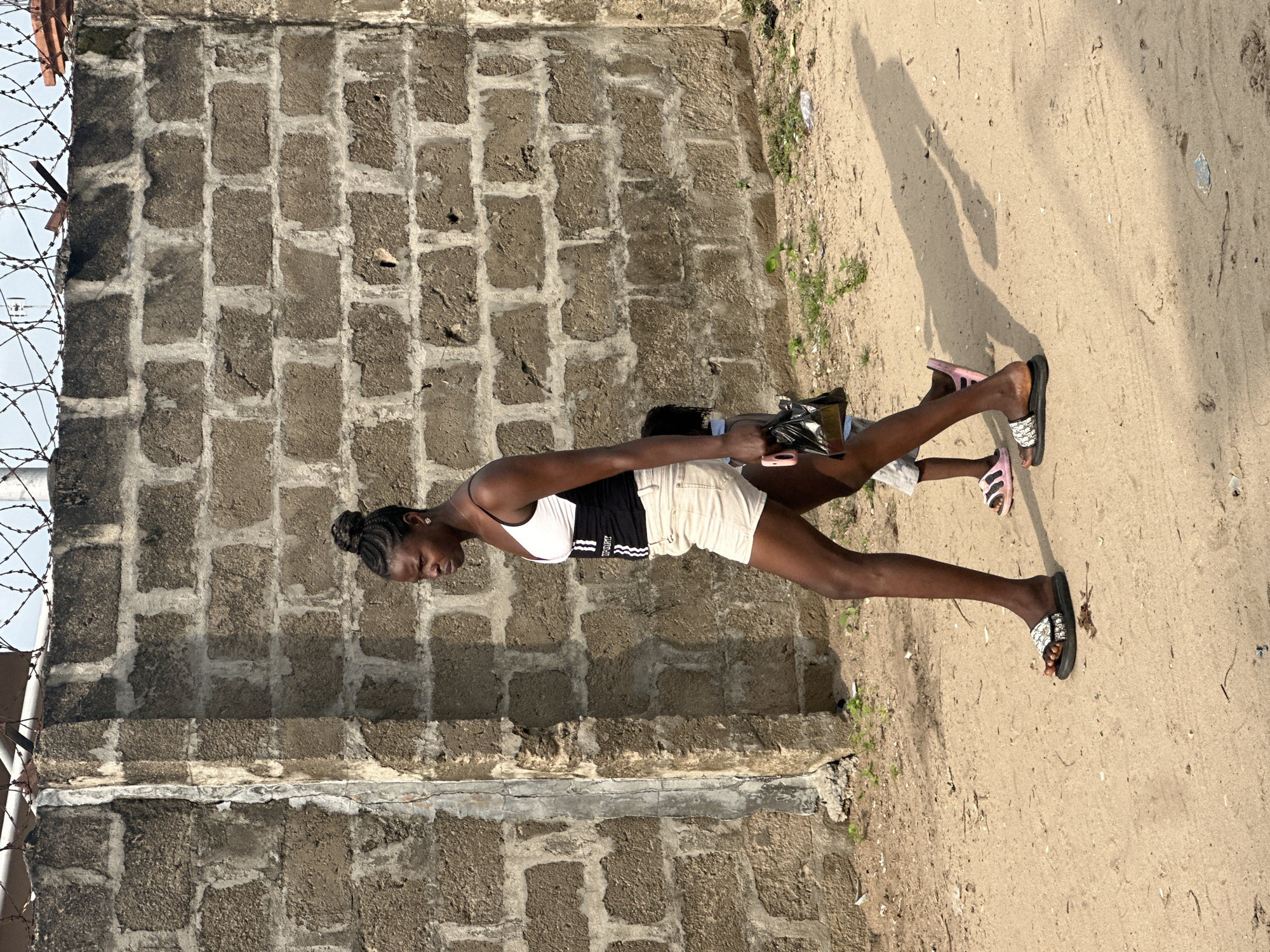In Badagry, Nigeria, a young woman with her hair in a bun walks along a sandy path beside a weathered brick wall topped with barbed wire. She wears a white and black athletic-style tank top and light-colored shorts, carrying a black purse in her right hand. Beside her, a child wearing a grey shirt and pink slide sandals walks, their face obscured. The sun casts long shadows on the ground.
