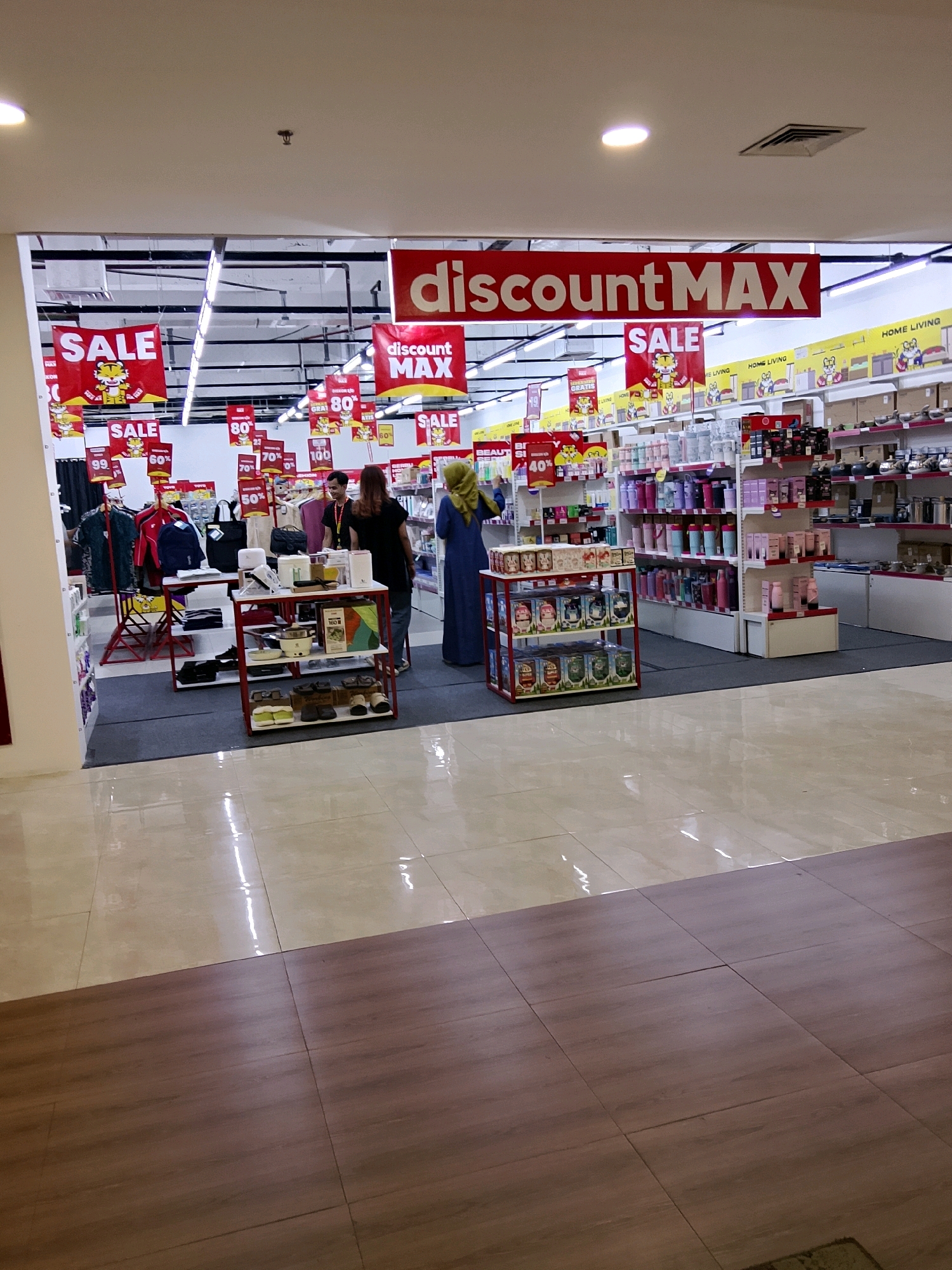 The image shows the entrance to a "discountMAX" store, likely a supermarket or department store, located in Cirebon, Indonesia. The entrance is framed by a white archway. Inside, there are various sale signs, mostly red with white lettering, indicating discounts such as "SALE", "80%", "70%", "50%", and "40%". Display shelves are filled with a variety of merchandise, including clothing on racks, and what appear to be household items, personal care products, and packaged goods. Two women and one man are visible within the store. One woman, wearing a blue hijab and blue dress, is browsing items on a shelf. Another woman, with brown hair and wearing a black shirt and blue jeans, is standing near a display table. A man, wearing a dark t-shirt, is standing behind a counter or display. The floor inside the store is a combination of light-colored tiled flooring and darker wood-like flooring in the foreground. Fluorescent lighting is visible on the ceiling, illuminating the store.