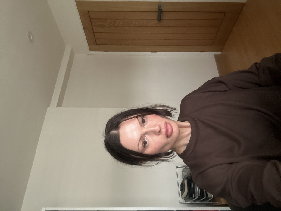 A female individual is depicted indoors in Tunbridge Wells, United Kingdom. She has dark, medium-length hair parted centrally, pulled back from her face, with some strands falling forward. Her facial features include pronounced cheekbones and full lips. She wears a dark brown, long-sleeved crew-neck top. The subject is positioned centrally, facing directly towards the camera, with her upper torso and shoulders visible.
The background consists of plain, light-colored walls and a light-colored wooden floor. To the right, a light brown wooden door with vertical paneling is present, featuring a horizontal, dark-colored handle or latch. A recessed light fixture is visible on the ceiling in the upper right. To the left of the subject, a portion of a metal multi-tiered trolley or shelf unit is visible, containing stacked items. The lighting appears to be ambient, illuminating the room evenly.