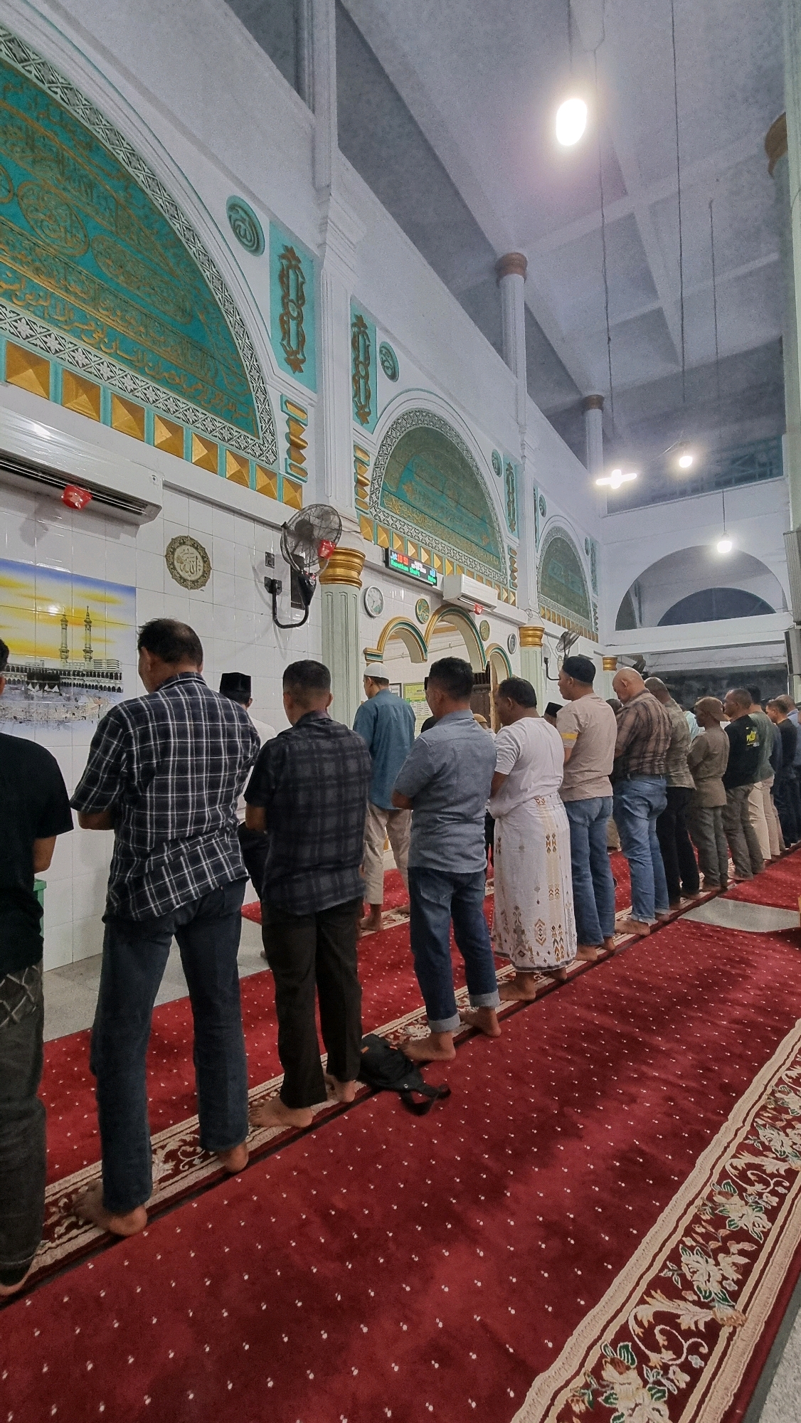 A group of men stand in prayer on a red carpet inside a mosque. The men are dressed in various clothing, including shirts, pants, and traditional Islamic attire. The mosque features white walls adorned with decorative green and gold arches, as well as Arabic calligraphy. A painting depicting the Kaaba in Mecca hangs on the left wall. The lighting suggests it is daytime, with natural light coming from windows or openings at the top of the structure, supplemented by artificial lights. The scene appears to be a religious gathering in Puloara, Indonesia.