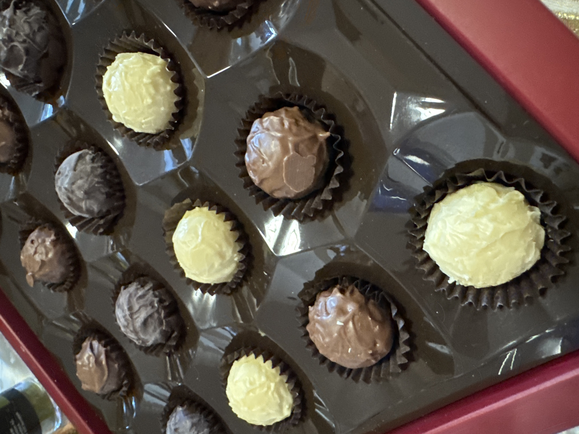 A close-up shot shows an opened box of chocolate truffles. The box is dark brown, and some of the truffles sit in black paper wrappers. The truffles vary in color from light brown and dark brown, with some having a pale yellow tint. The truffles are nestled in individual compartments within the box. The lid of the box is red. The scene takes place indoors. Location information (Sofia, Bulgaria) is not discernible from the image.