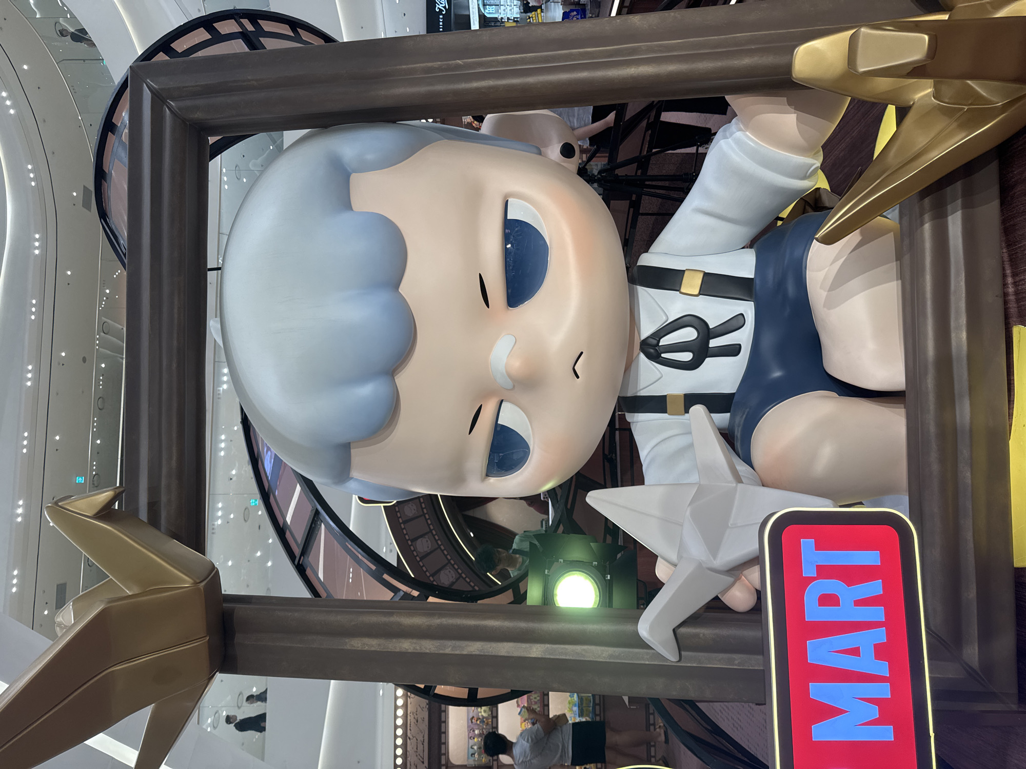 This media file shows a large, stylized cartoon sculpture of a young person with pale skin, light blue hair, and large blue eyes. The sculpture is framed by a thick, dark brown border. The figure is wearing a white shirt with suspenders, dark blue shorts, and white socks. They are holding a white, star-shaped object in their left hand.
The sculpture appears to be part of a display or advertisement, as indicated by a vertical sign with red background and blue text that reads "MART". The setting is an indoor environment, likely a shopping mall or commercial space, with modern architecture featuring white curved walls and ceiling lights. In the background, there are glimpses of escalators, other displays, and people walking around. One person is visible in the foreground, standing and looking down, and other people are visible in the background, appearing to be shopping.
The image is captured from a low angle, looking up at the sculpture. The lighting appears to be artificial, consistent with an indoor setting. There are no clear indications of the time of day or weather conditions. The overall impression is that of a vibrant, commercial display designed to attract attention.