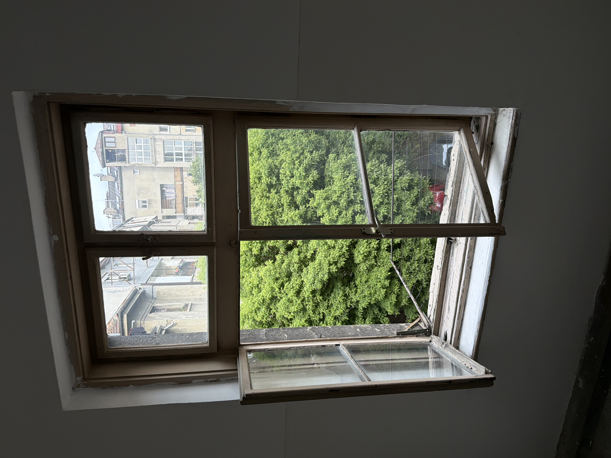 The image depicts an open window in a building located in Belgrade, Serbia. The window, framed in wood and painted white, has a section that swings outward, allowing a view of lush green trees. Through the closed section of the window, buildings typical of Belgrade's architecture are visible. The interior walls surrounding the window are painted a neutral gray. The overall lighting suggests it is daytime.