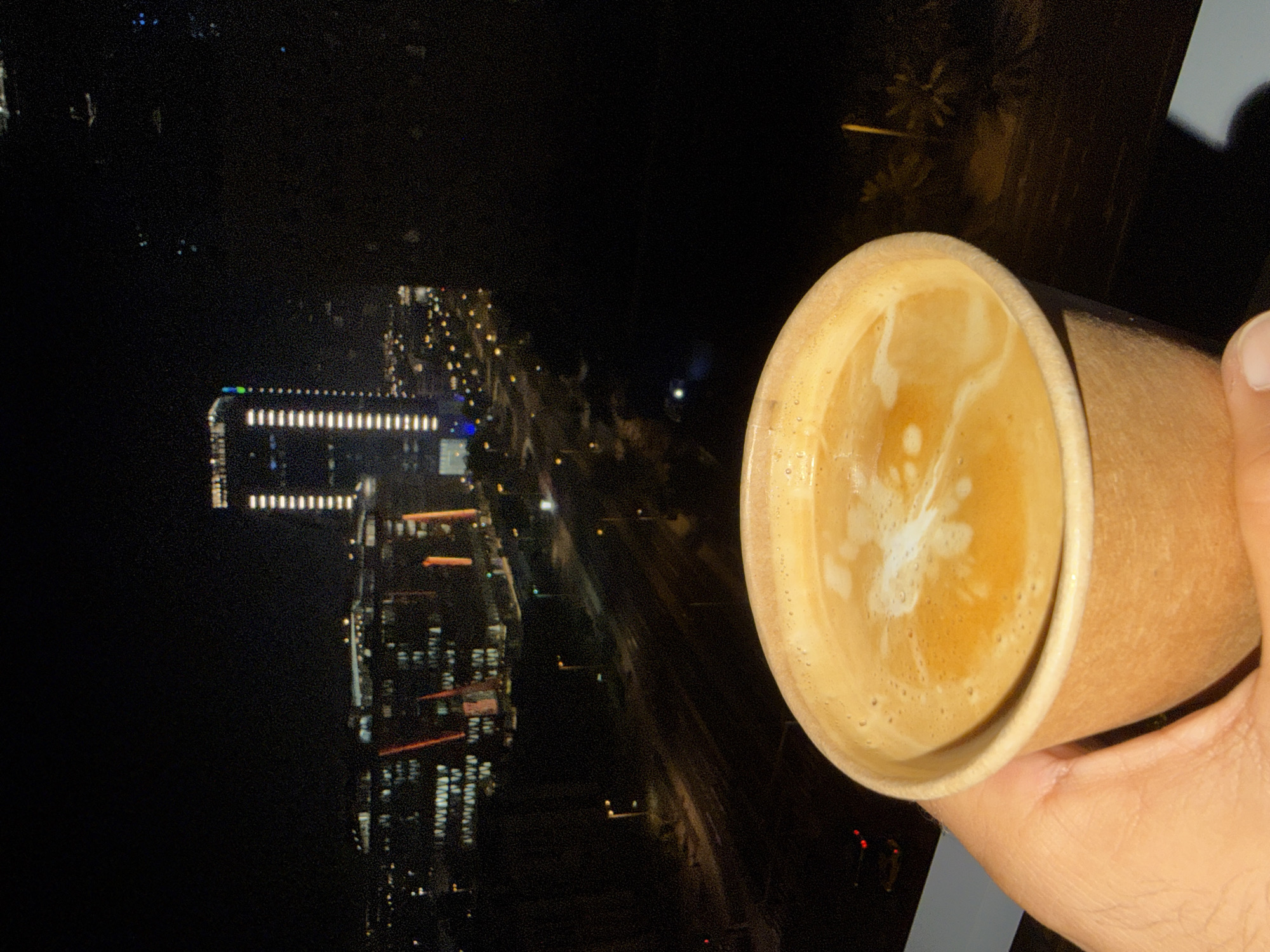 A hand holds a light brown paper cup of coffee with foam, in the foreground. Behind the cup, a city skyline is visible at night. Tall buildings with illuminated windows and streetlights create a dense urban landscape. The scene appears to be viewed from an elevated position, likely a window.