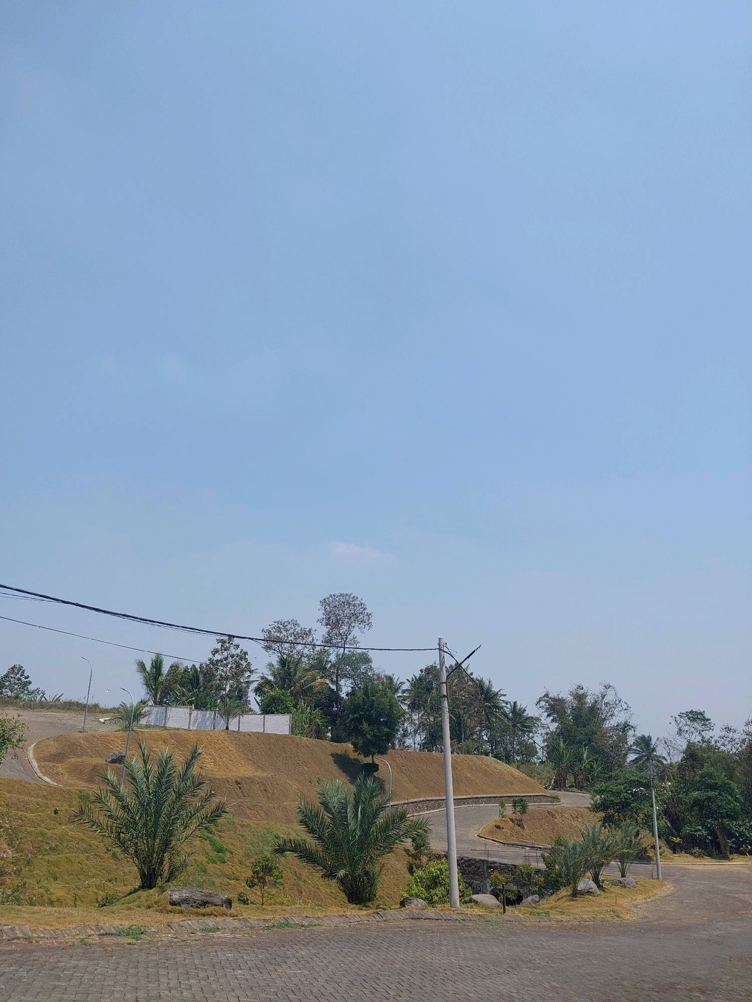 The image depicts a gently sloping, grassy hill with a paved pathway winding through it. Several palm trees and other vegetation are scattered across the landscape. A white fence is visible at the top of the hill, and power lines stretch across the sky. The sky is clear and blue. The foreground is paved with cobblestones. The scene appears to be a park or a residential area in Malang, Indonesia. The lighting suggests it is daytime, likely late morning or early afternoon due to the bright, uniform illumination. There are no people visible in the image. The overall impression is one of a well-maintained outdoor space.