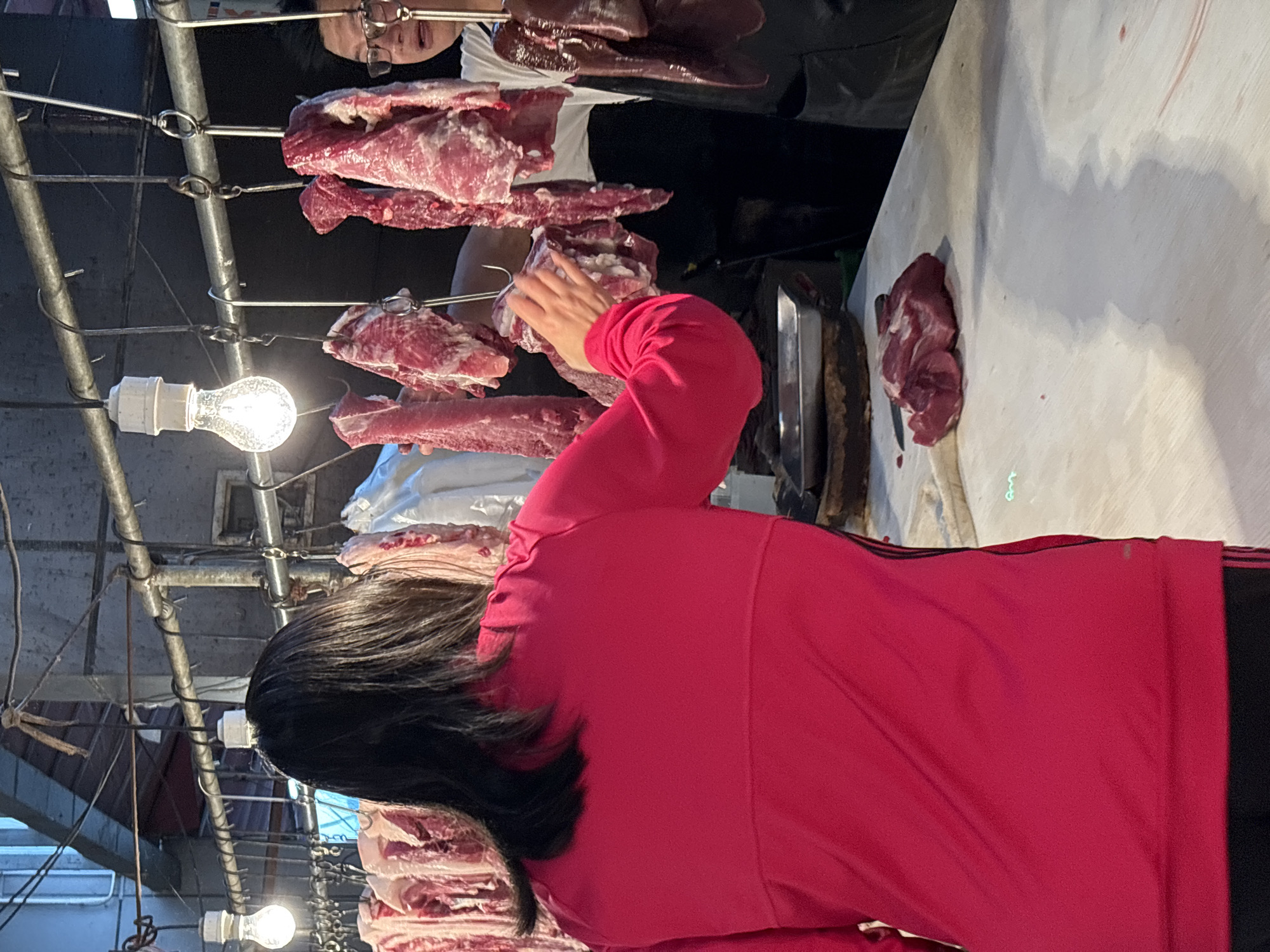 The image shows a meat stall, presumably in Jiangjin, China, based on the prompt. A person in a pink top appears to be selecting pieces of meat hanging on metal hooks. Other people are also present in the background. The stall has a white table, and some meat is placed on it. Overhead is a network of metal bars and wires supporting the hanging meat and light bulbs. The environment appears to be indoors.