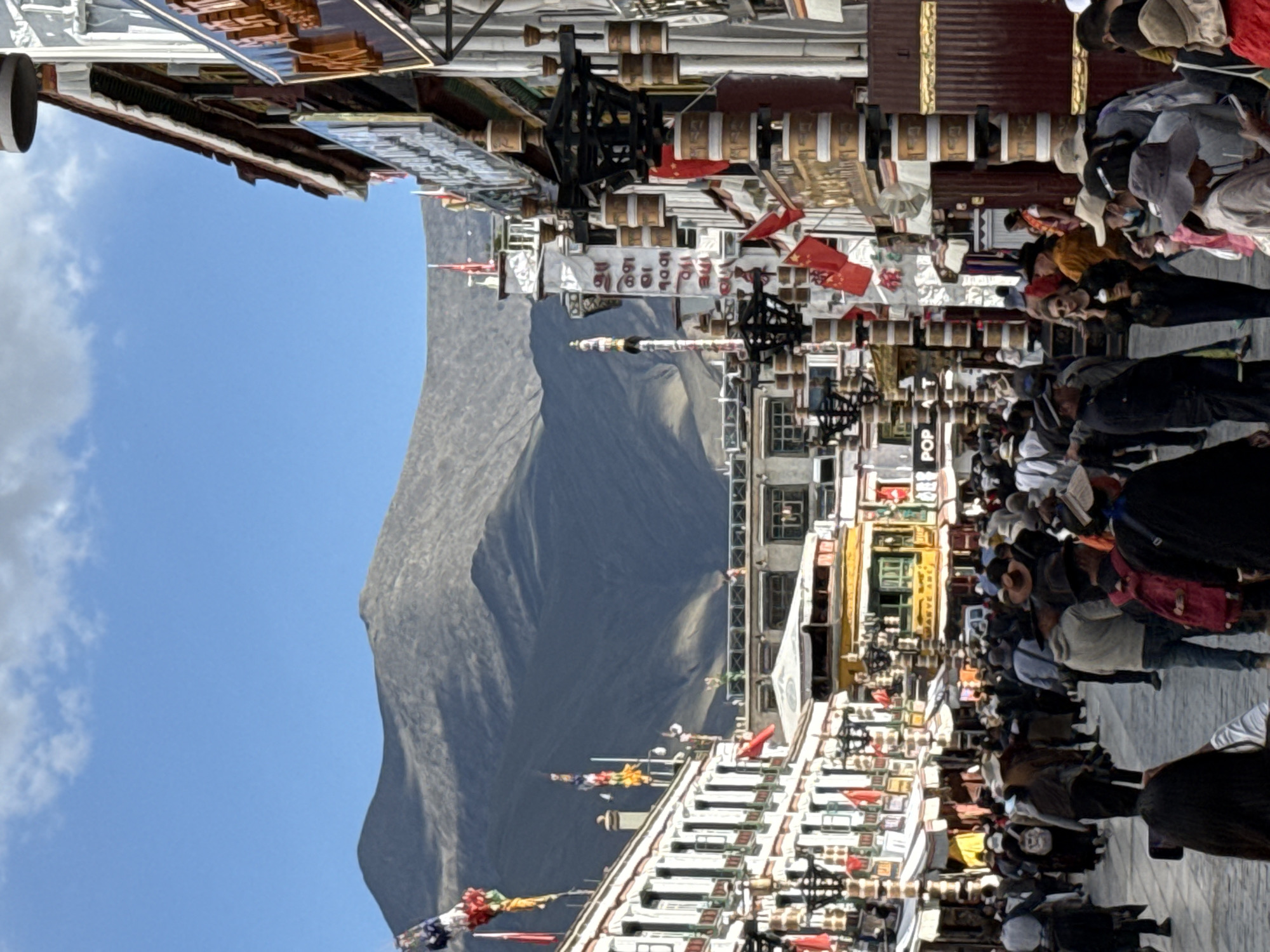 The image, rotated 90 degrees clockwise for proper viewing, depicts a bustling street scene set against a majestic mountain backdrop under a clear blue sky with scattered clouds. In the foreground and midground, numerous people are seen walking and standing on a street lined with multi-story buildings. The architecture suggests a Tibetan cultural influence, featuring traditional elements and many large cylindrical prayer wheels affixed to the facades of the buildings. Red Chinese flags are prominently displayed, hanging from poles and strung across the street, alongside colorful prayer flags (lungta).
The street appears to be a vibrant public space, possibly a market or a path leading to a religious site, given the concentration of people and cultural symbols. In the far background, a large, barren, and imposing mountain dominates the horizon, suggesting a high-altitude, mountainous region. The time of day appears to be bright daylight.
Visible text includes Tibetan script on a white banner stretched across the street, which appears to read "བཀྲ་ཤིས་བདེ་ལེགས" (Tashi Delek), a common Tibetan greeting. Another partially visible sign on a building reads "POP". The overall atmosphere is lively and crowded, indicative of a busy day in an area rich with cultural and natural significance in Chengguan District, China.