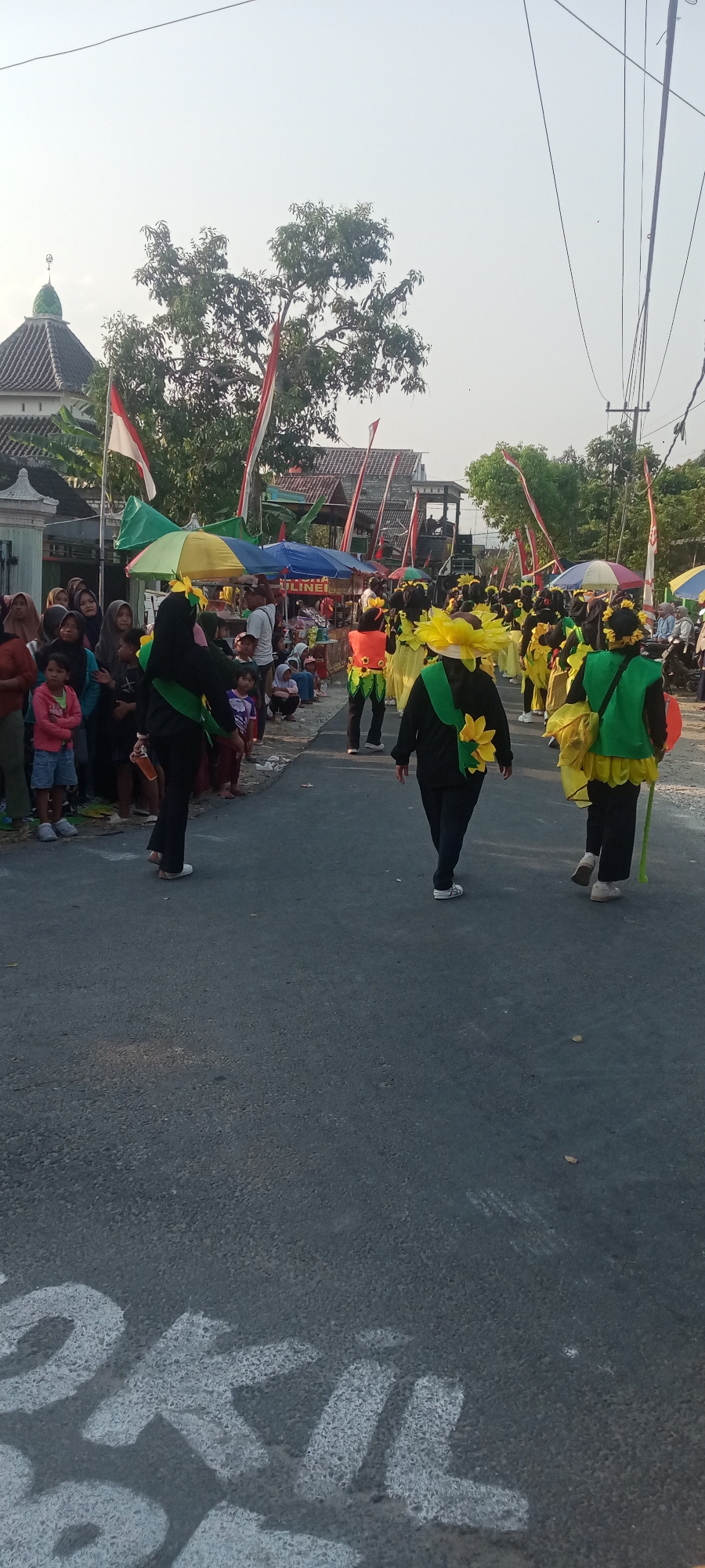 This is a vertical, eye-level, full-shot photograph taken in Soko, Indonesia. The scene depicts a street parade, likely during a festive occasion, with a crowd of spectators gathered on the left side of the road. The spectators are a mix of adults and children, some dressed in traditional Islamic clothing, and others in casual attire.
The parade participants are dressed in vibrant costumes, predominantly yellow and green, with large, sunflower-like embellishments. They are walking down the asphalt road. Some participants are wearing black headscarves and black pants, adorned with green vests and yellow "sunflower" accessories on their chests and heads. Others are wearing yellow dresses and green vests. One participant in the foreground is wearing an orange vest over a yellow outfit.
On the left side of the road, there are several tents and stalls set up, suggesting a temporary market or food vendors. Indonesian flags, red and white, are visible along the street, indicating a national celebration. In the background, a mosque with a green dome can be seen. The sky is bright and clear, suggesting daytime. White chalk markings are visible on the road in the foreground, with some letters partially visible, possibly part of a race or event marking. The overall atmosphere appears lively and community-oriented.