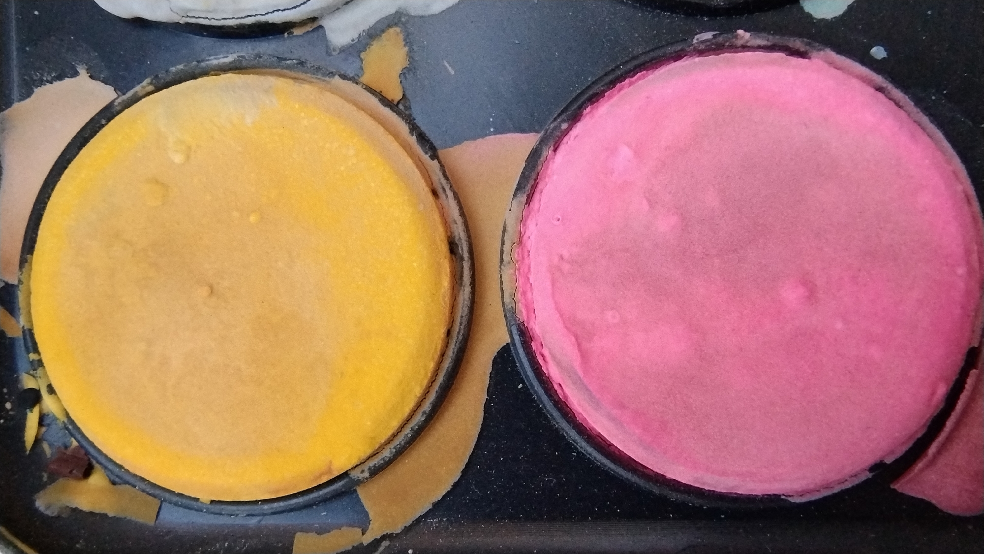 This media file is a close-up shot of a palette of face paints. The subjects are two circular pans of face paint, one yellow and one pink. They are nestled within a black palette, which appears to be made of plastic. The pans are full of what looks like pressed powder makeup. The yellow paint is a bright, sunny hue, while the pink paint is a soft, pastel shade.
The paints are round and sit snugly in their compartments within the palette. The surface of the yellow paint has a slightly uneven texture with small bumps or indentations. The pink paint also has a somewhat textured surface, with a few tiny raised dots visible. The edges of the pans are lined with a dark rim.
Around the pans, there are pieces of what appears to be paper or cardboard peeling away from the palette. These pieces are a light tan or beige color and add a somewhat worn or used appearance to the overall scene. The background is dark, likely the rest of the palette, with some fine dust or debris visible.
No people or specific setting are visible, suggesting the focus is solely on the makeup itself. There is no visible text in the image. The lighting appears to be overhead and somewhat diffused, creating soft shadows. The overall impression is one of a well-used but still functional makeup palette, ready for artistic application.