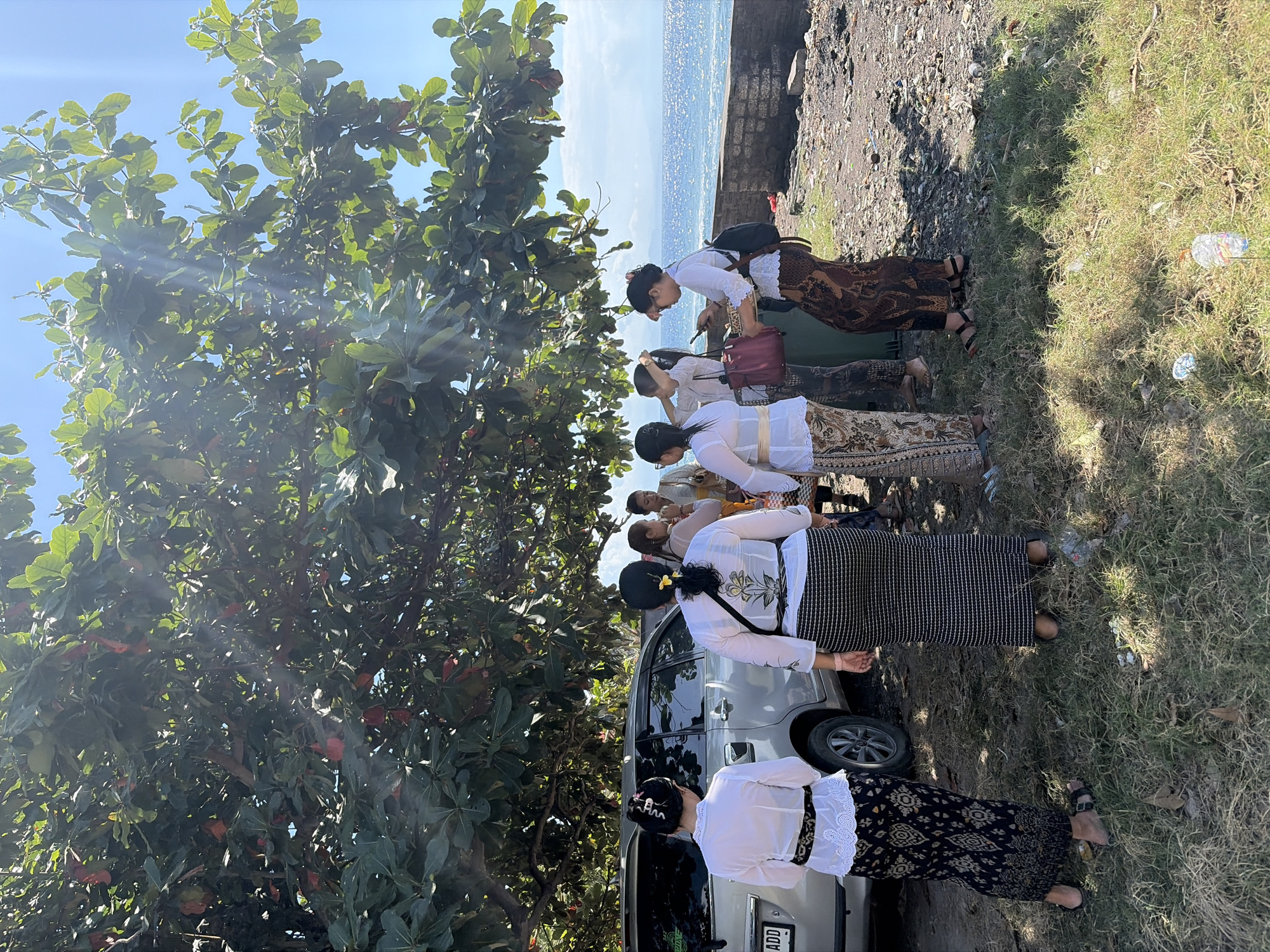 A group of women in traditional attire stand near a silver SUV by the ocean on Nusa Penida, Indonesia. The women are dressed in white tops and long, patterned skirts. Some are holding bags and looking at their phones. The SUV is parked on a dirt and gravel area with dry grass. In the background, the turquoise ocean sparkles under a bright blue sky, with a low stone wall separating the shore from the land. Sunlight filters through the leaves of a large tree overhead, casting dappled shadows on the ground.