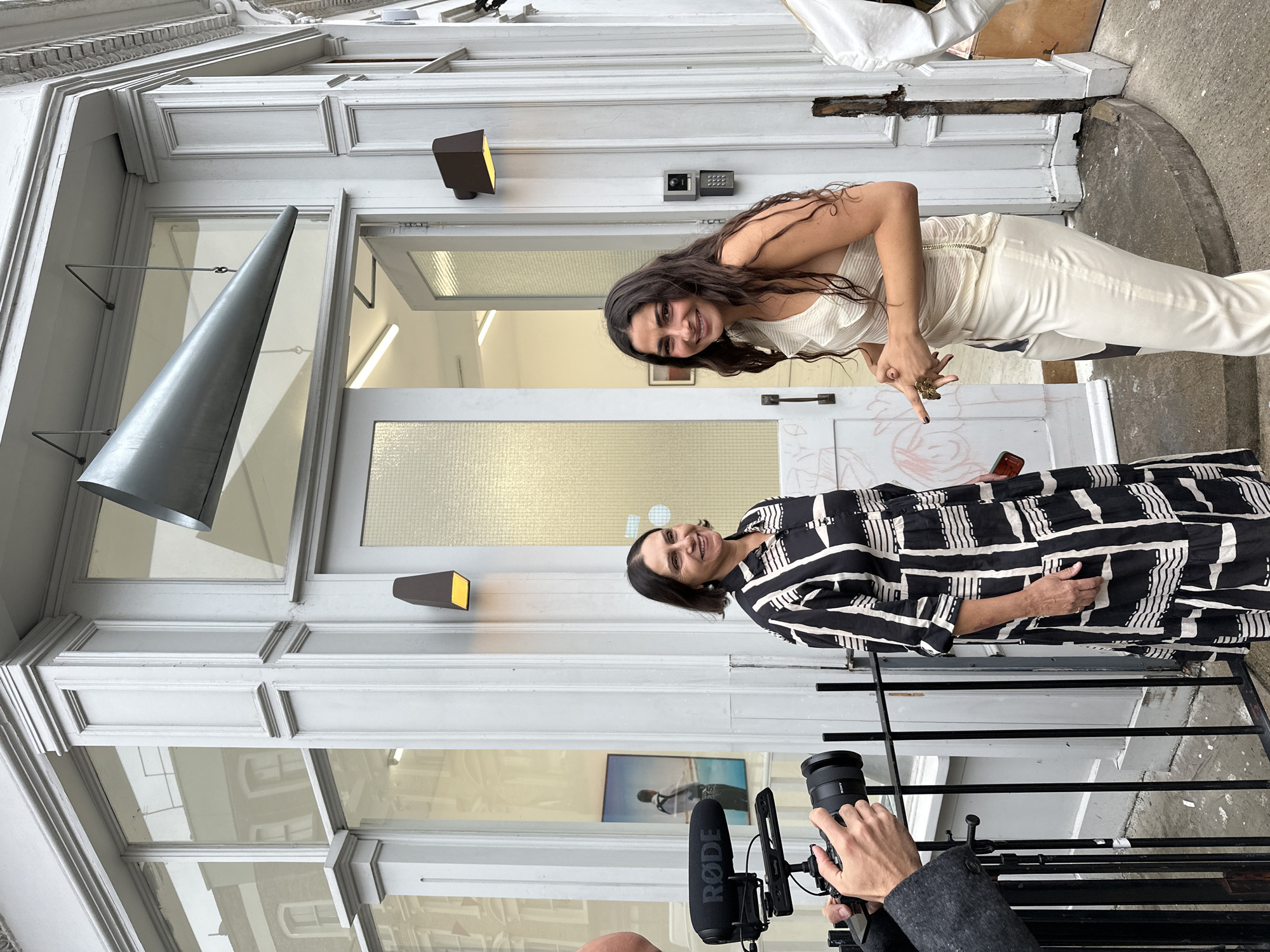 Two women stand outside a white building with large glass windows and a grey door. The woman on the left wears a black and white patterned dress and the woman on the right wears a white asymmetrical top and matching trousers. A black camera with a Rode microphone is being held by an unseen person in the foreground. The building appears to be a shop or gallery, with what looks like artwork visible through the window. A large metallic cone-shaped fixture hangs above the entrance. The scene suggests an event or opening is taking place. The building's architecture and the overall aesthetic suggest it is located in a city like London, United Kingdom, possibly in an area known for its art galleries or boutique shops.