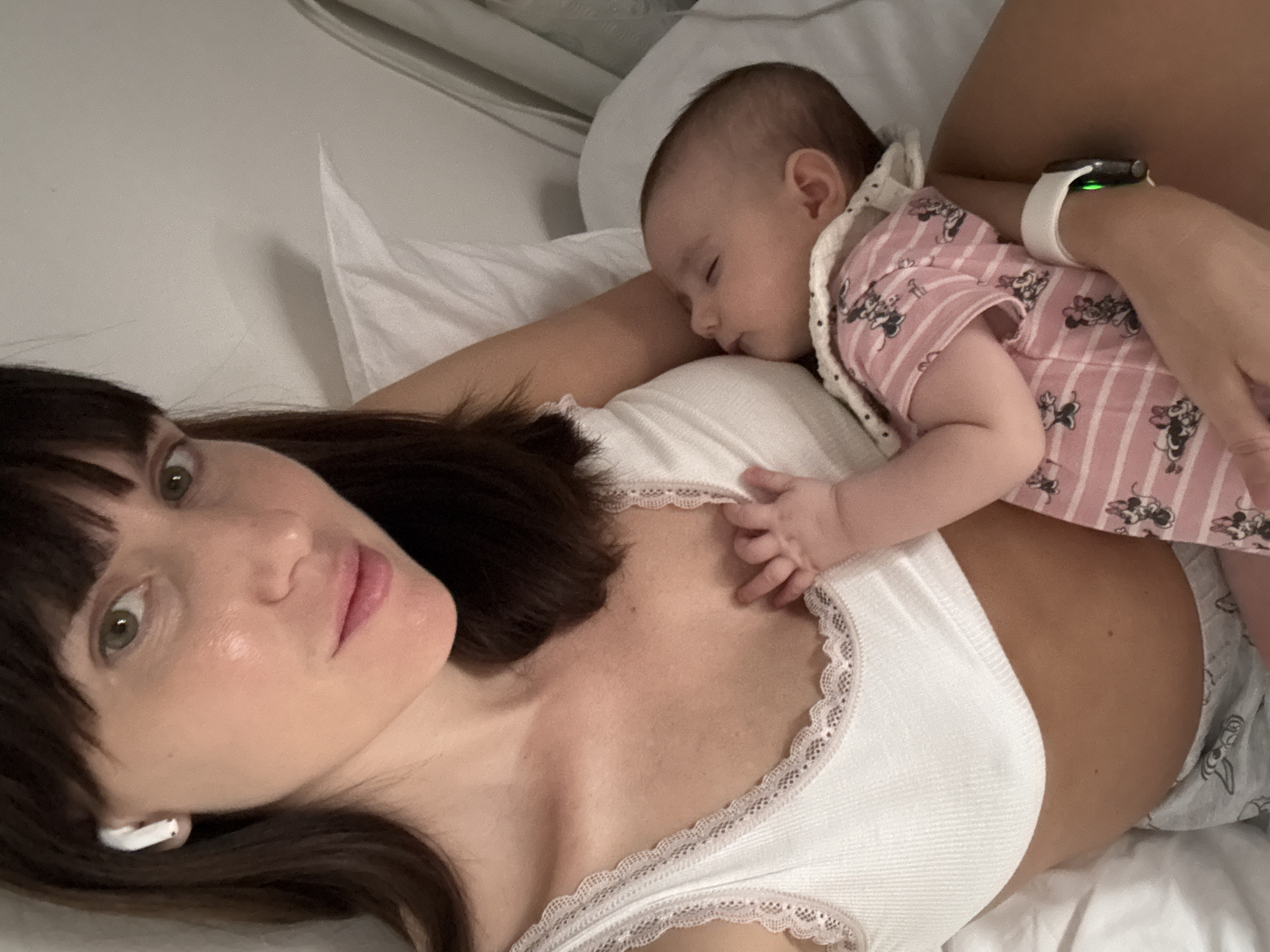 A woman with dark hair and bangs, wearing a white lace-trimmed tank top and grey shorts with cartoon characters, holds a sleeping baby on her chest. The baby is wearing a pink and white striped outfit with cartoon images and a white bib. The woman has an earbud in her left ear and a smartwatch on her right wrist. They are lying down, possibly in a bed, with white pillows and sheets visible. The scene is dimly lit. The location is not specified in the image or its context.