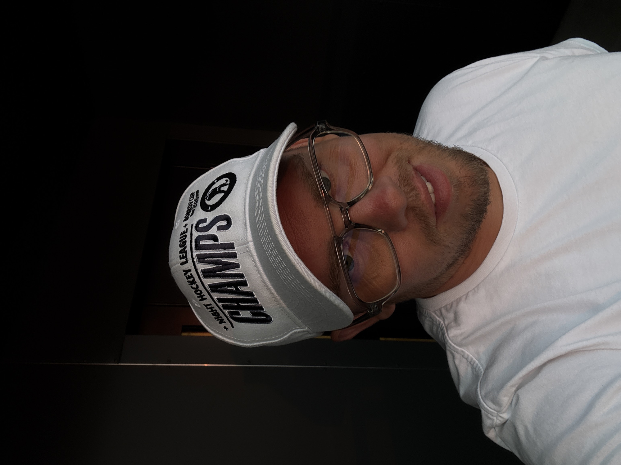 The media file is a close-up, sideways selfie of a man wearing a white t-shirt and a white visor. The visor has black text and a black logo. The visible text on the visor reads "CHAMPS" prominently, with smaller text above and below it. The smaller text above appears to be "LEAGUE," and the text below reads "NIGHT HOCKEY." The logo on the visor is a circular emblem with a silhouette of a hockey player. The man has short brown hair and is wearing clear-rimmed glasses. He has a light beard and is smiling with his mouth slightly open, showing his teeth. The background is dark and out of focus, providing no discernible location cues other than the text on the visor. The lighting suggests an indoor setting, possibly at night, given the dark background. The subject's expression appears happy and relaxed. The presence of "CHAMPS" and "NIGHT HOCKEY" on the visor suggests the man may be celebrating a hockey victory or is a participant in a hockey league. The context provided indicates the location is Sochi, Russia, but this cannot be visually confirmed from the image itself.