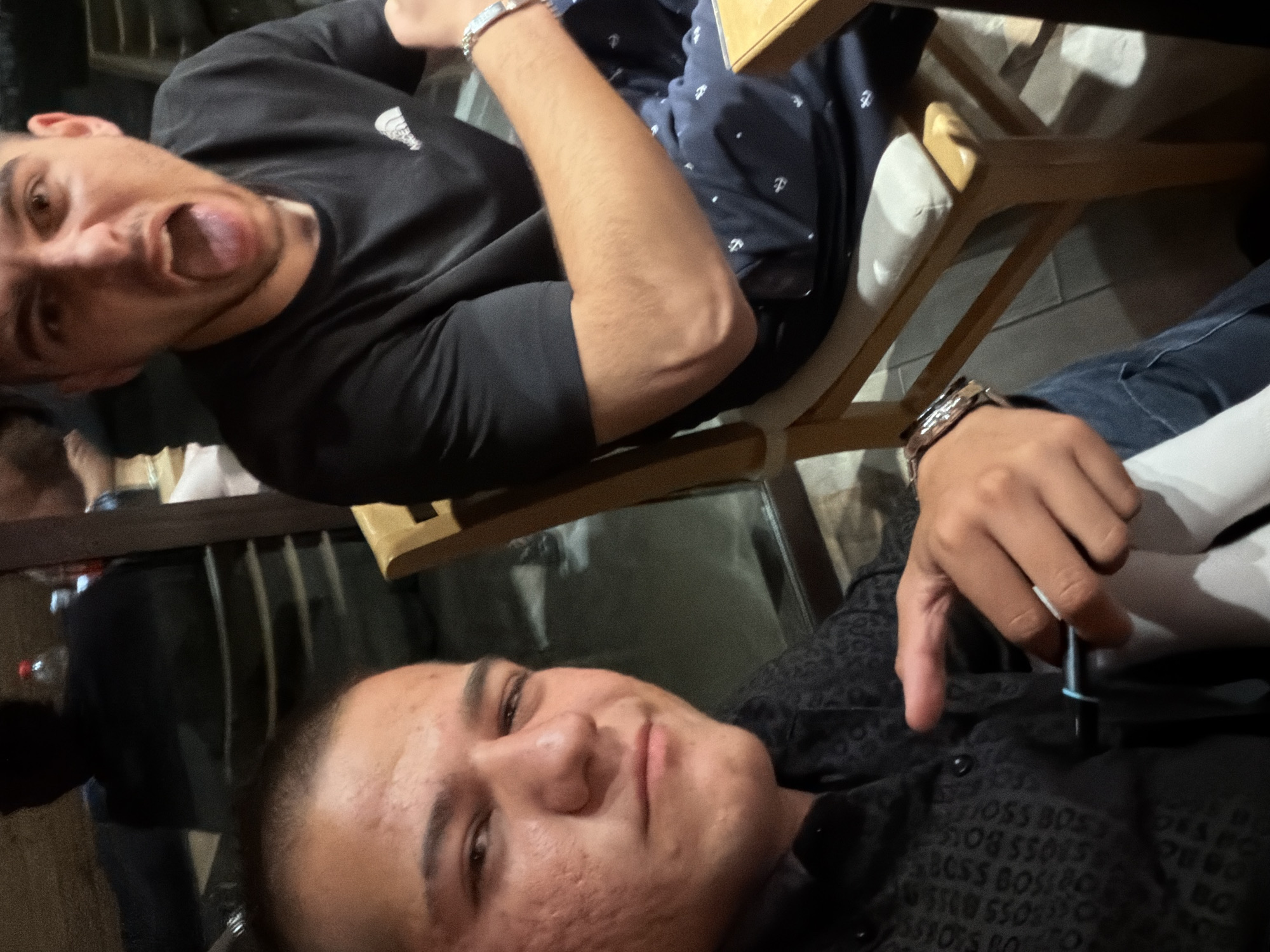 Two young men are the primary subjects in this image, taken in what appears to be a restaurant in Svejen, Bulgaria. The man on the left is in the foreground, taking a selfie. He has short, dark hair and is wearing a dark-colored shirt with a repeating "BOSS" logo pattern. His facial skin shows some texture. He is holding a pen-like object in his right hand, near his chest.
The second man is seated to the right and slightly behind the first. He is wearing a black t-shirt and has his mouth wide open, with his tongue visible, suggesting he is making a playful or surprised expression. He has a watch on his left wrist. He is seated in a wooden chair with a light-colored cushion.
The background suggests an indoor setting, possibly a restaurant with reflective surfaces, like windows or a dark partition, showing blurred reflections of people and possibly bottles on a shelf. The lighting is dim, indicating an evening or indoor environment. The overall impression is a candid snapshot of friends interacting.