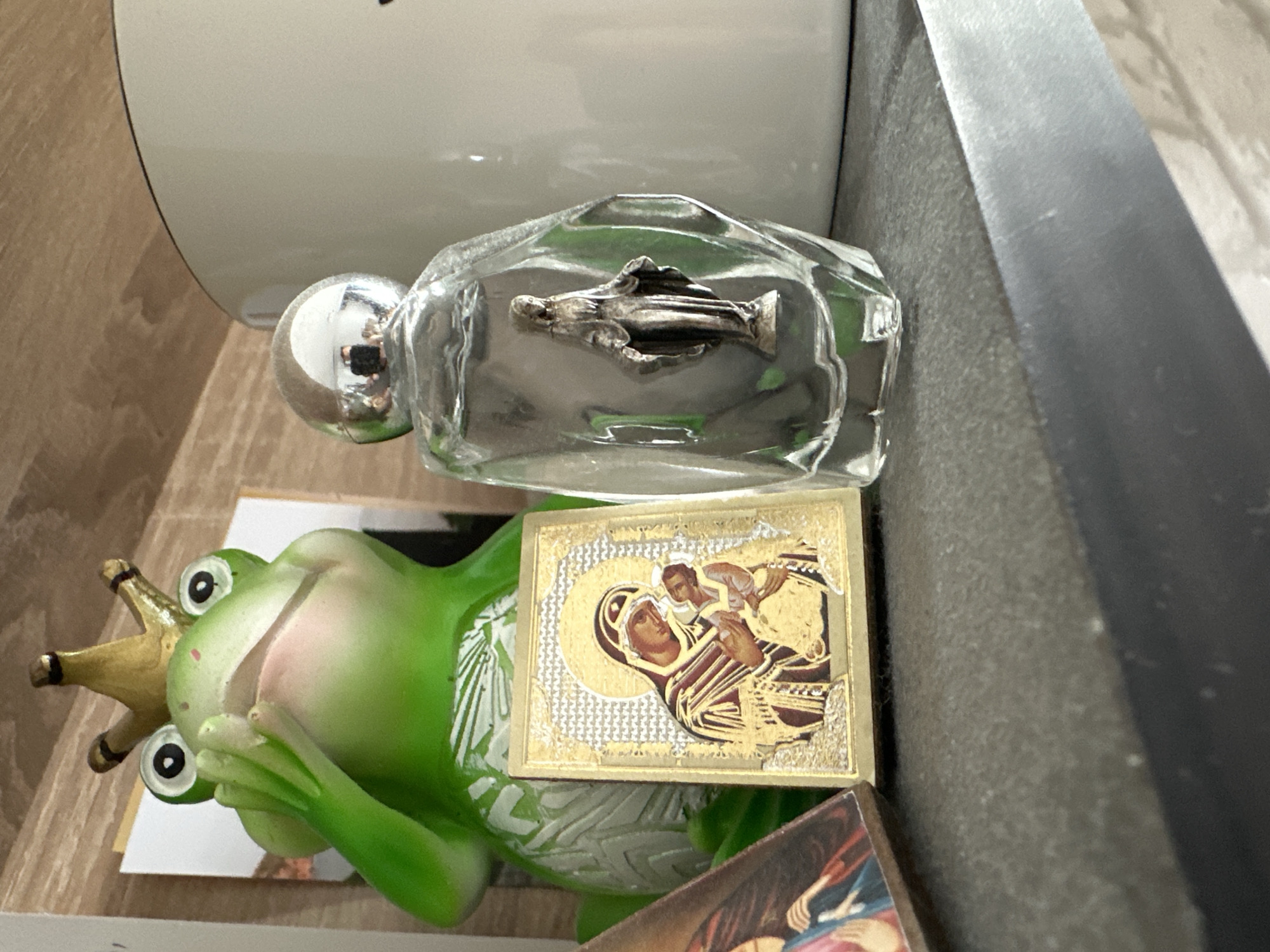 The image captures a close-up, low-angle view of an indoor arrangement of decorative and religious items on a dark gray, textured surface in Unknown, Unknown. A bright green frog figurine, wearing a gold crown and covering its mouth with its right hand, is positioned on the left. The frog displays a white, leafy pattern on its lower body. Adjacent to the frog, a small, rectangular religious icon with a gold frame and textured background depicts the Virgin Mary holding baby Jesus, rendered primarily in gold and dark brown tones. To the immediate left of this icon, a corner of another small, brown-edged icon displaying figures in blue and red is partially visible. To the right of the gold icon, a clear, faceted glass bottle with a rounded silver cap contains a small, silver statuette of the Virgin Mary with outstretched arms. The silver cap exhibits a clear reflection of a human figure holding a camera. Behind the items, a large, light cream-colored ceramic mug is partially visible on the right, and a wooden paneling background with a distinct grain pattern occupies the upper portion of the frame. A white card is positioned behind the frog. The items rest on a flat, dark gray surface with a slight beveled edge in the foreground.