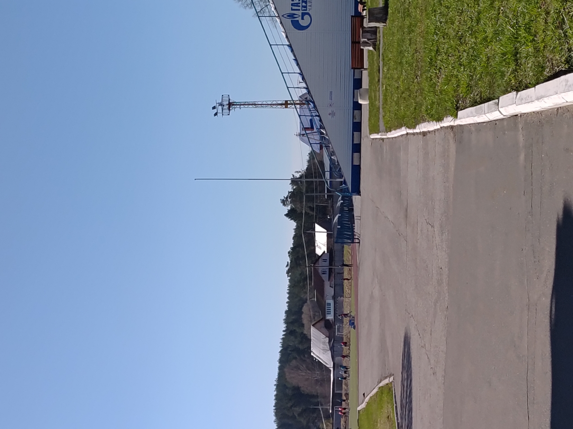 The image depicts an outdoor scene in Chaikovsky, Russia. A grey asphalt road runs along the right side, bordered by a concrete curb and green grass. Adjacent to the grass is a white and blue building with the Gazprom logo. In the background, there are trees and houses. A metal tower stands near the building. The sky is clear and blue. People are faintly visible in the distance along the buildings on the road.