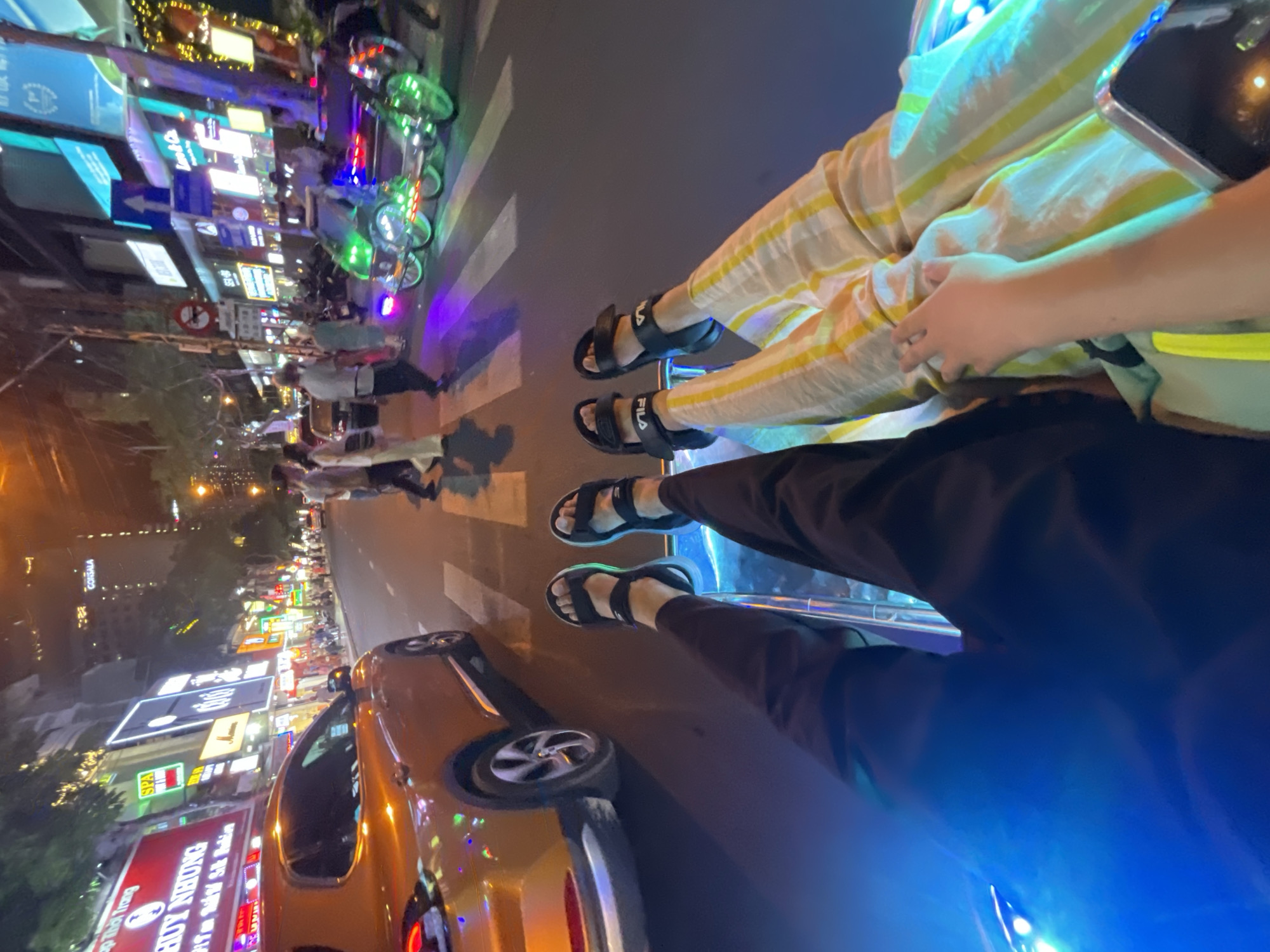 Captured from the perspective of an individual riding a vehicle on a city street in Nha Trang, Vietnam, the image reveals a vibrant urban scene at night. The legs and feet of two passengers are visible, one wearing yellow and white striped pants and black sandals, the other clad in dark pants and similar sandals. A gold-colored vehicle, possibly a car, is partially visible in the lower-left corner of the frame. The street is lined with businesses displaying illuminated signage, including what appears to be a spa and the word "HUY NHUNG". Pedestrians are seen walking along the crosswalk. Additional vehicles, including three-wheeled vehicles with prominent green neon lights, are also present. The time of day appears to be evening or nighttime, evidenced by the artificial lighting illuminating the city.