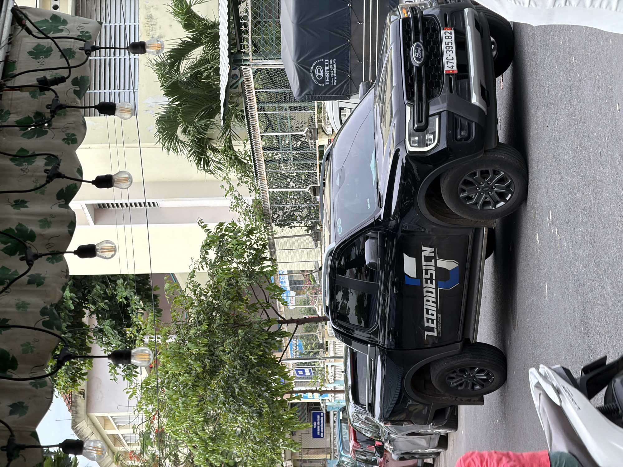 This image captures a daytime urban street scene in Nha Trang Ward, Vietnam. The prominent subject is a black Ford pickup truck, likely a Ranger model, parked on the side of a paved road. The truck features a custom white and blue decal on its side doors that reads "LEGIADESION" and "BAUERILISING." Its front license plate displays "47C-395.82," and the "Ford" logo is visible on the grille.
To the left, a string of decorative Edison-style light bulbs hangs beneath a patterned awning adorned with a green leaf motif, suggesting an outdoor cafe or shop entrance. Beyond this, multi-story buildings with windows and balconies line the street. Various green trees and foliage are interspersed throughout the scene, adding natural elements.
In the background, a large black tarp or cover, possibly over a truck or stall, displays advertising text including "CÔNG TY TNHH DAEHAN MOTORS," "TERACO," a "Hotline: 0902 409 909," and "www.daehan.vn." Further down the street, other vehicles are partially visible, including cars and a white scooter in the foreground. A small blue sign in the distance shows the Vietnamese text "YÊU CẦU." The lighting suggests it is daytime, possibly overcast, with no specific human activity clearly depicted, implying a moment of parked vehicles in a quiet street setting.