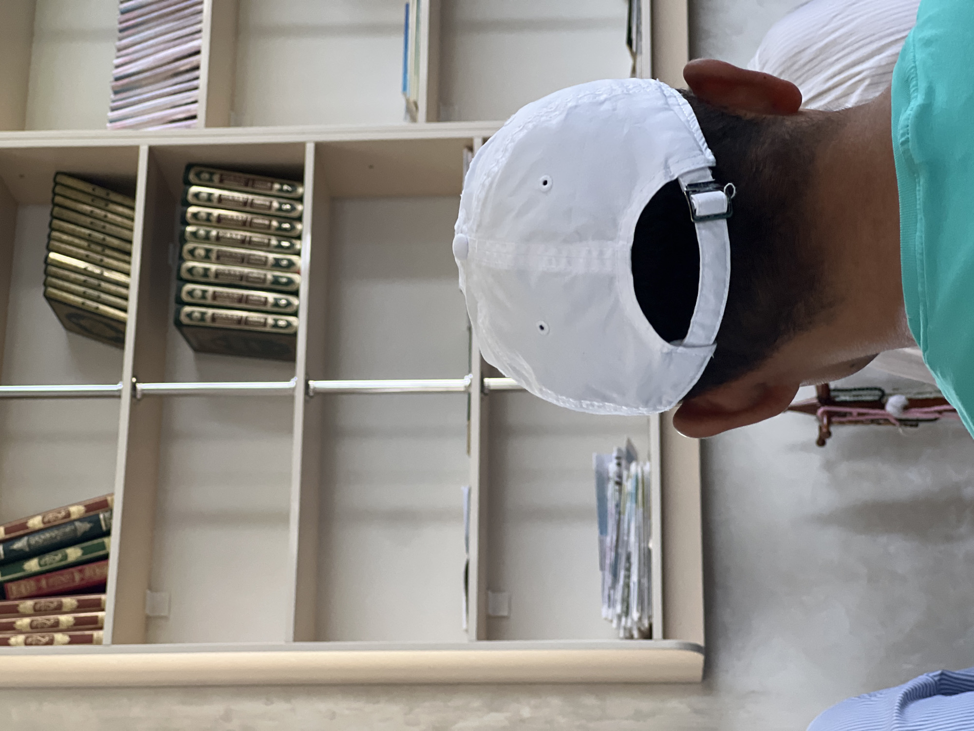 The image shows the back of a person wearing a white baseball cap and a teal t-shirt, facing away from the camera. Behind them is a white bookshelf with several shelves. The top shelf contains a row of books with gold lettering and dark covers, followed by a row of books with gold lettering and red and green covers. The second shelf from the top contains a row of books with gold lettering and dark green covers. The third shelf contains a stack of papers or thin books. The fourth shelf from the top contains a stack of papers or thin books. The right side of the bookshelf has more shelves with books. The background appears to be a wall with a textured pattern. The scene is indoors, and the lighting suggests it is daytime. The location is identified as Karamanas, Uzbekistan.