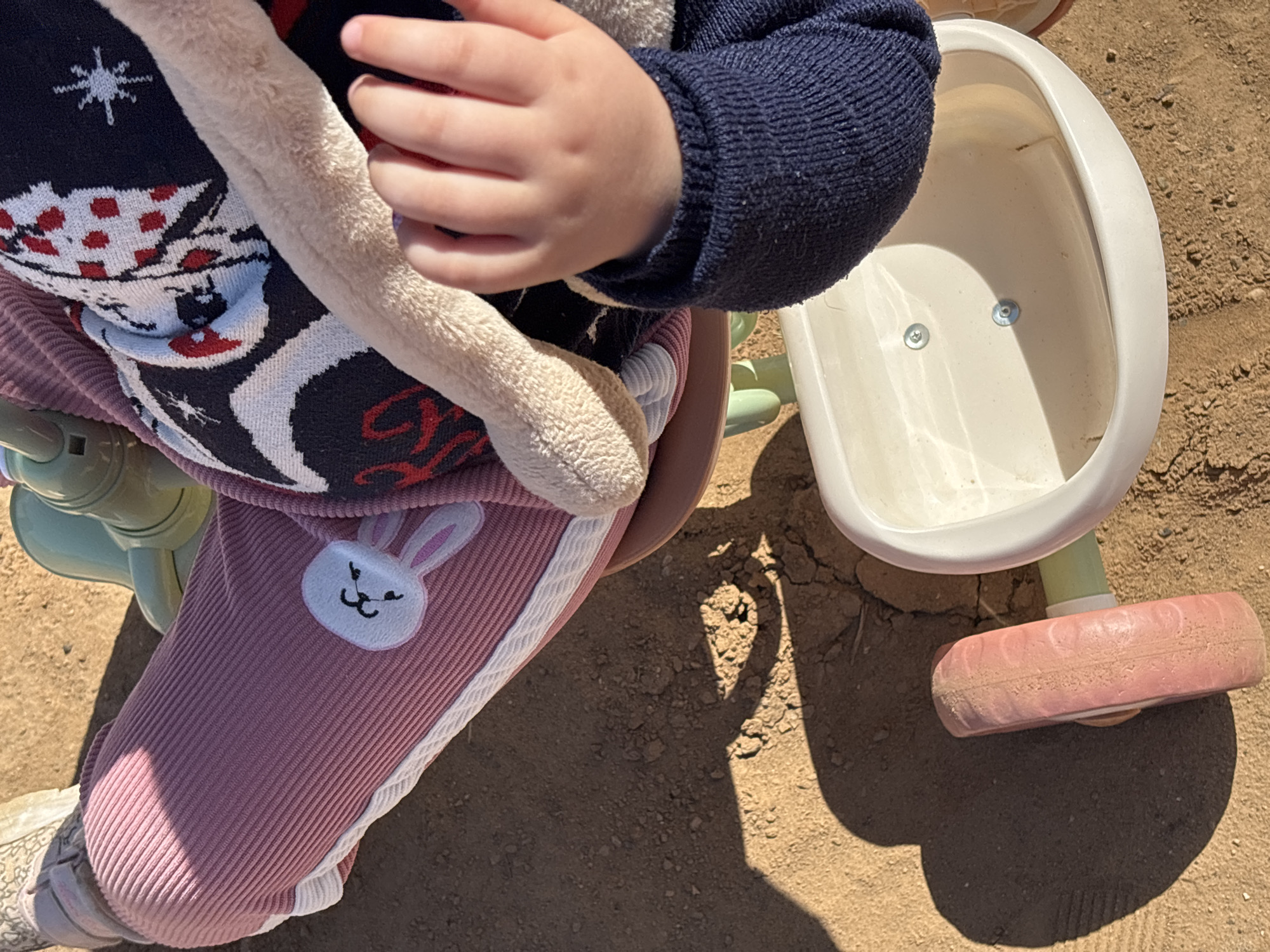 A toddler sits on a tricycle on a sunny day, likely in the city of Prikamskii, Russia. The tricycle has a white seat and a pink rear wheel. The toddler is wearing pink corduroy pants with a white stripe and a bunny applique, a dark blue sweater, and a patterned sweater over that. The toddler's hand is visible holding a stuffed animal. The ground is dry and cracked earth.